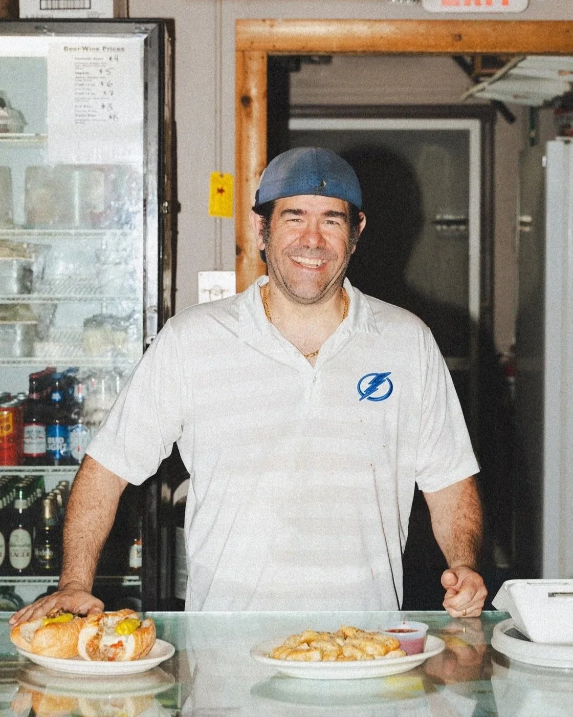 A man smiling behind a counter with hot dogs and pizza, wearing a white shirt with a blue lightning bolt logo and a blue cap, in a casual restaurant setting.