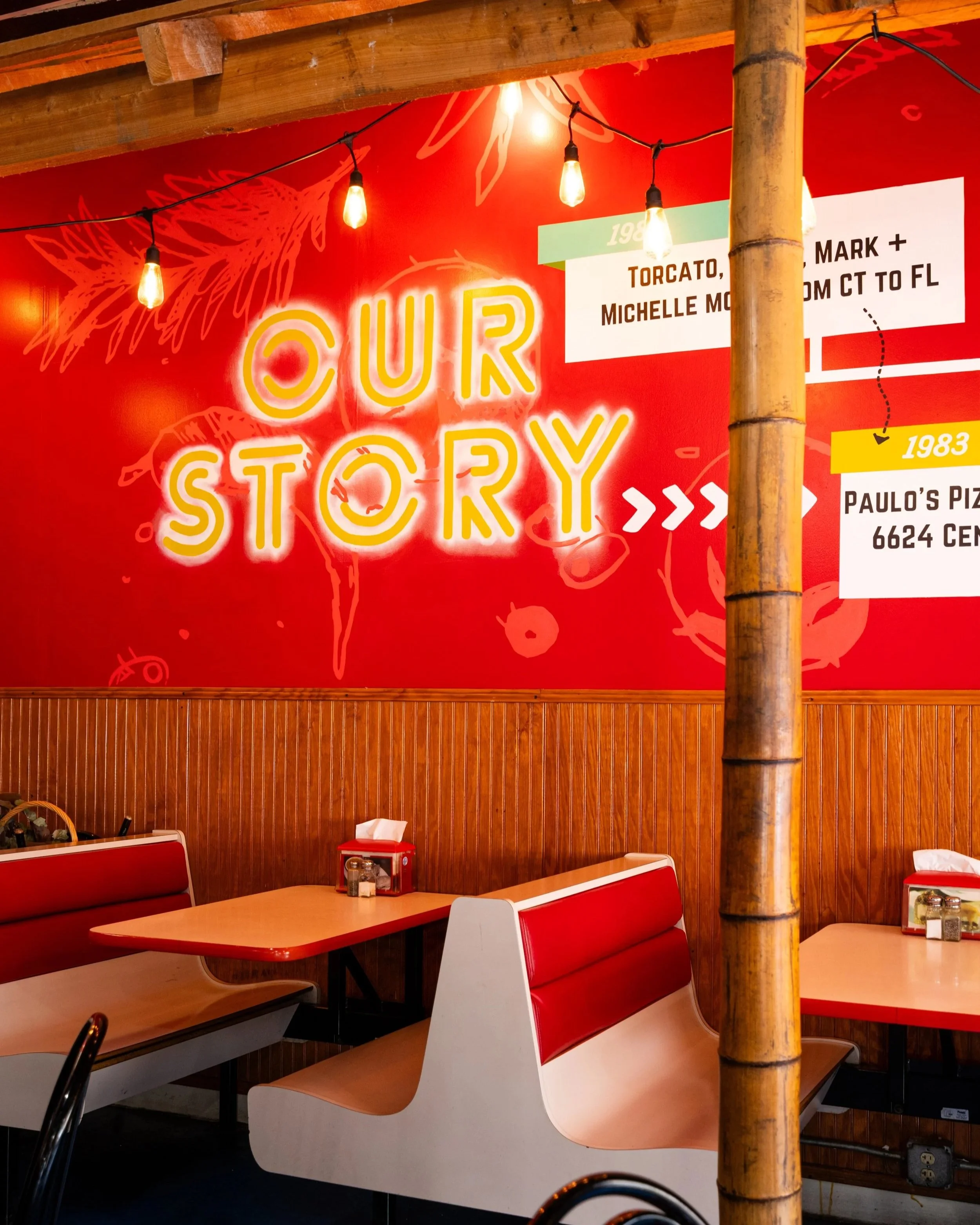 Indoor dining area of a restaurant with red and white booths, wood-paneled walls, a large bamboo pole, and warm lighting. Neon sign reading 'OUR STORY' is on the wall.