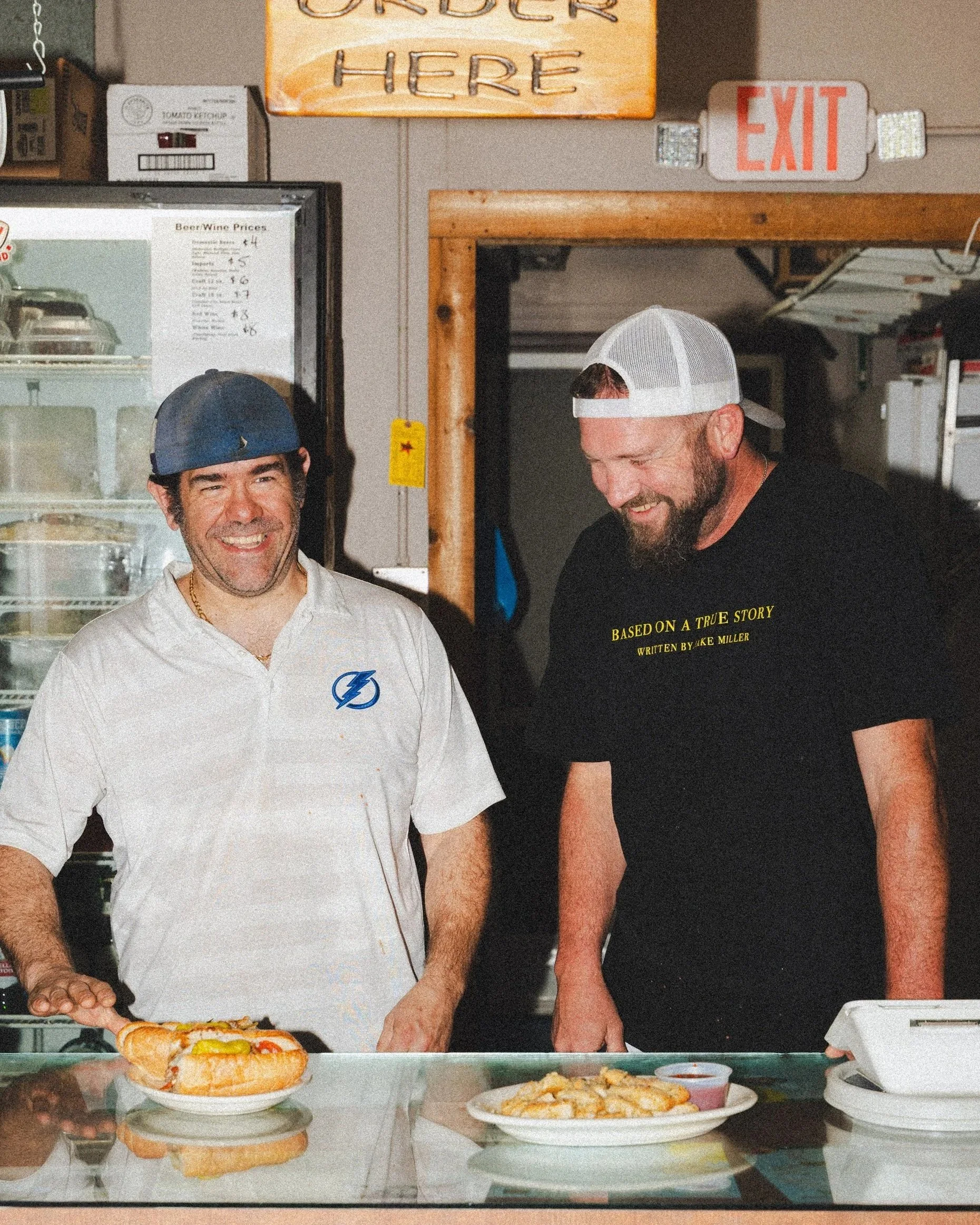 Two men working in a diner or sandwich shop. One is wearing a white chef's jacket and a blue cap, smiling, with a sandwich on a plate in front of him. The other man is wearing a black T-shirt and a white cap, preparing food. There are plates of food 