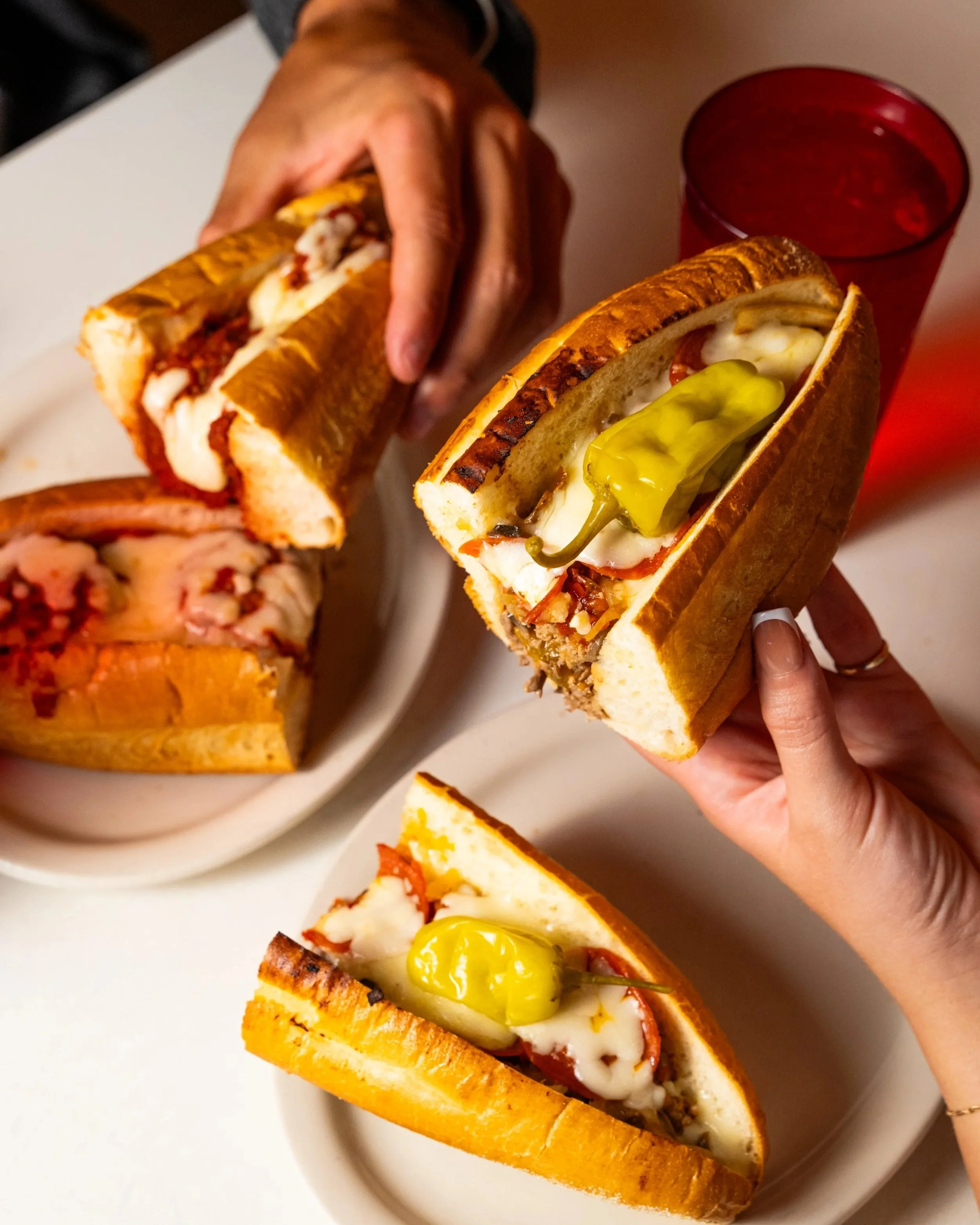 Close-up of two hot dogs with toppings, one with pickled pepper on a white plate, with a red plastic cup in the background.