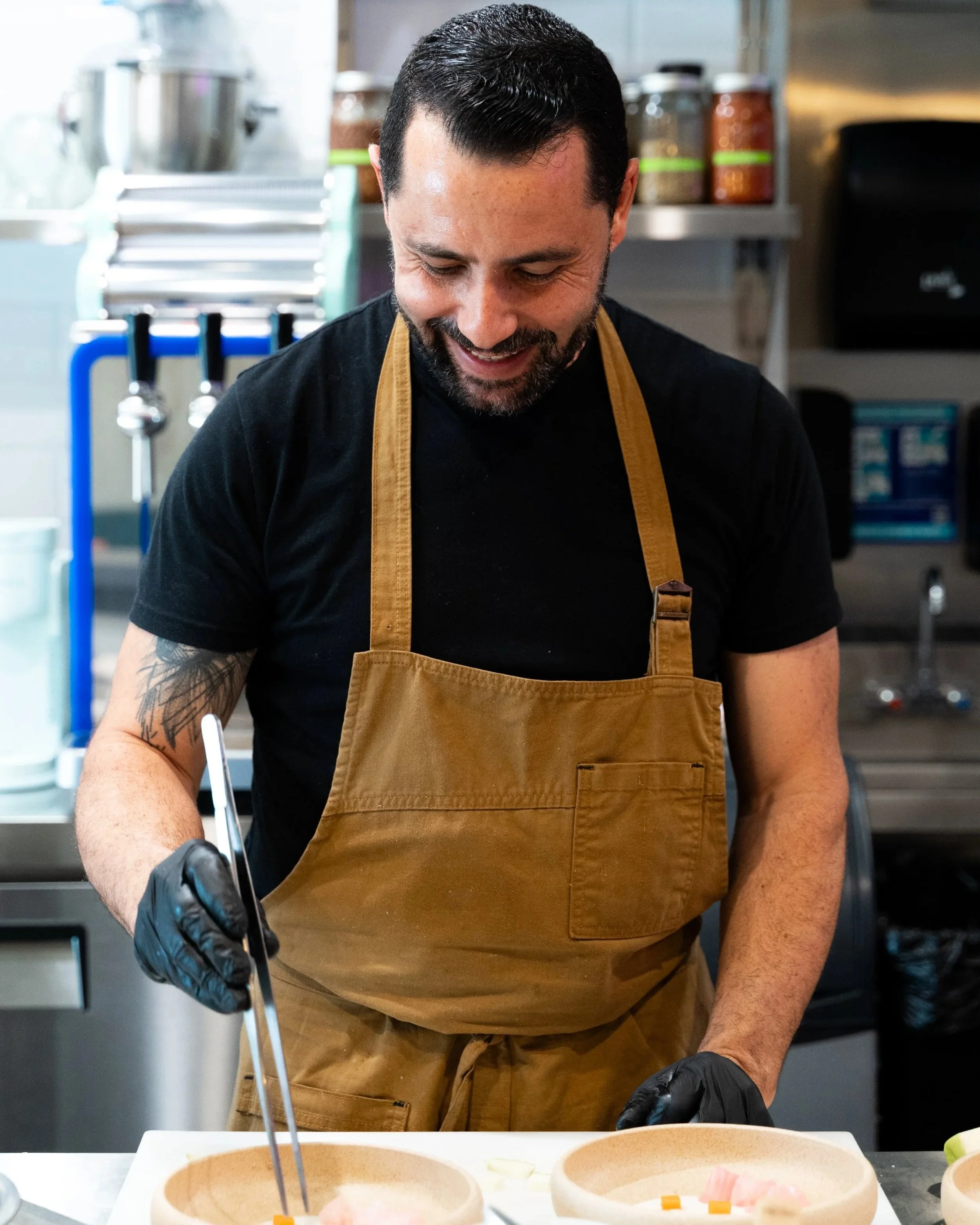 A man wearing a black t-shirt, a tan apron, and black gloves is smiling while preparing food in a commercial kitchen with jars of spices or ingredients in the background. Chef from Rakki in Tampa, Florida