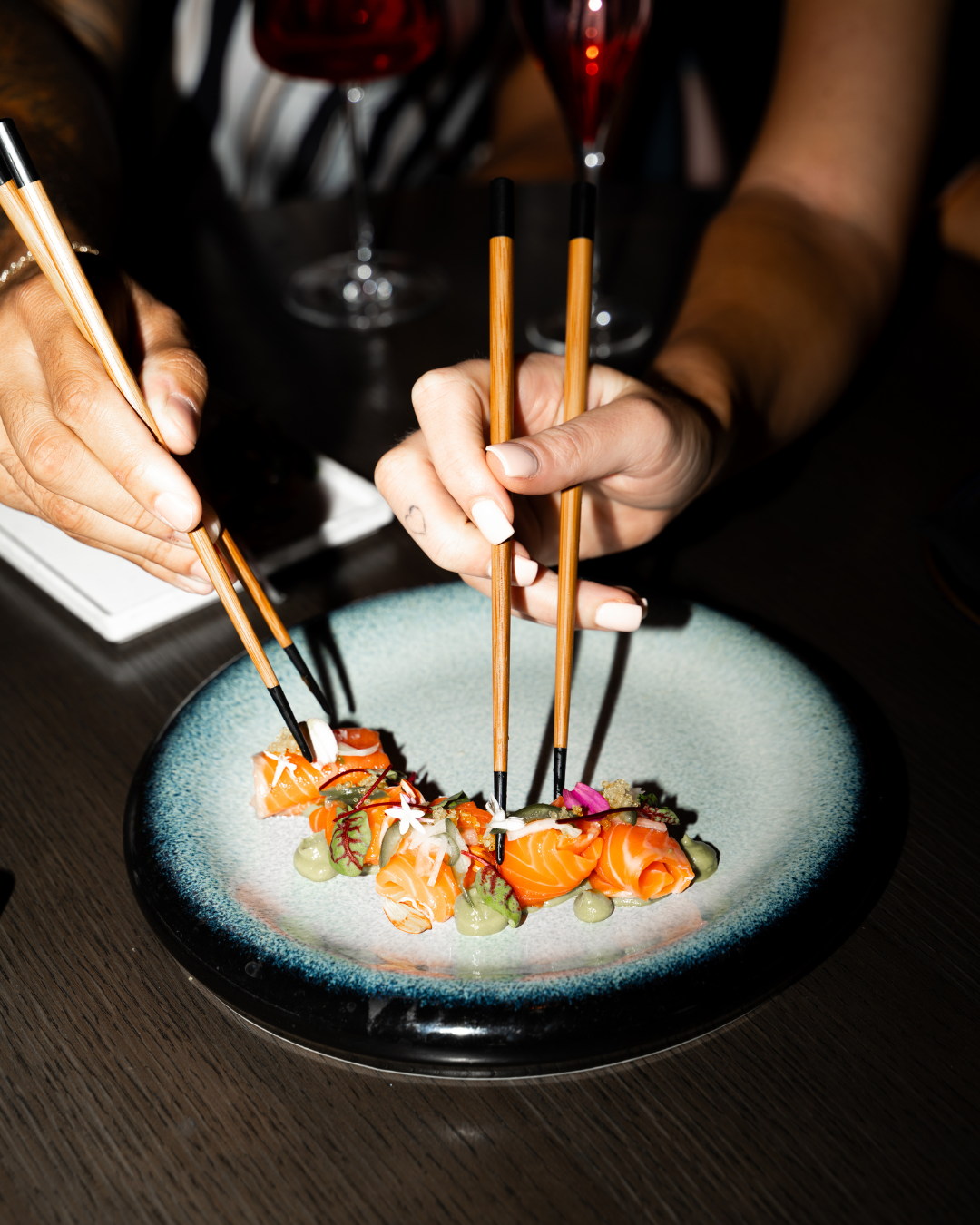 Hands using chopsticks to pick up sushi with salmon, garnishes, and microgreens on a blue ceramic plate, with glasses of red wine in the background. Ro Sushi Restaurant in Hyde Park. 