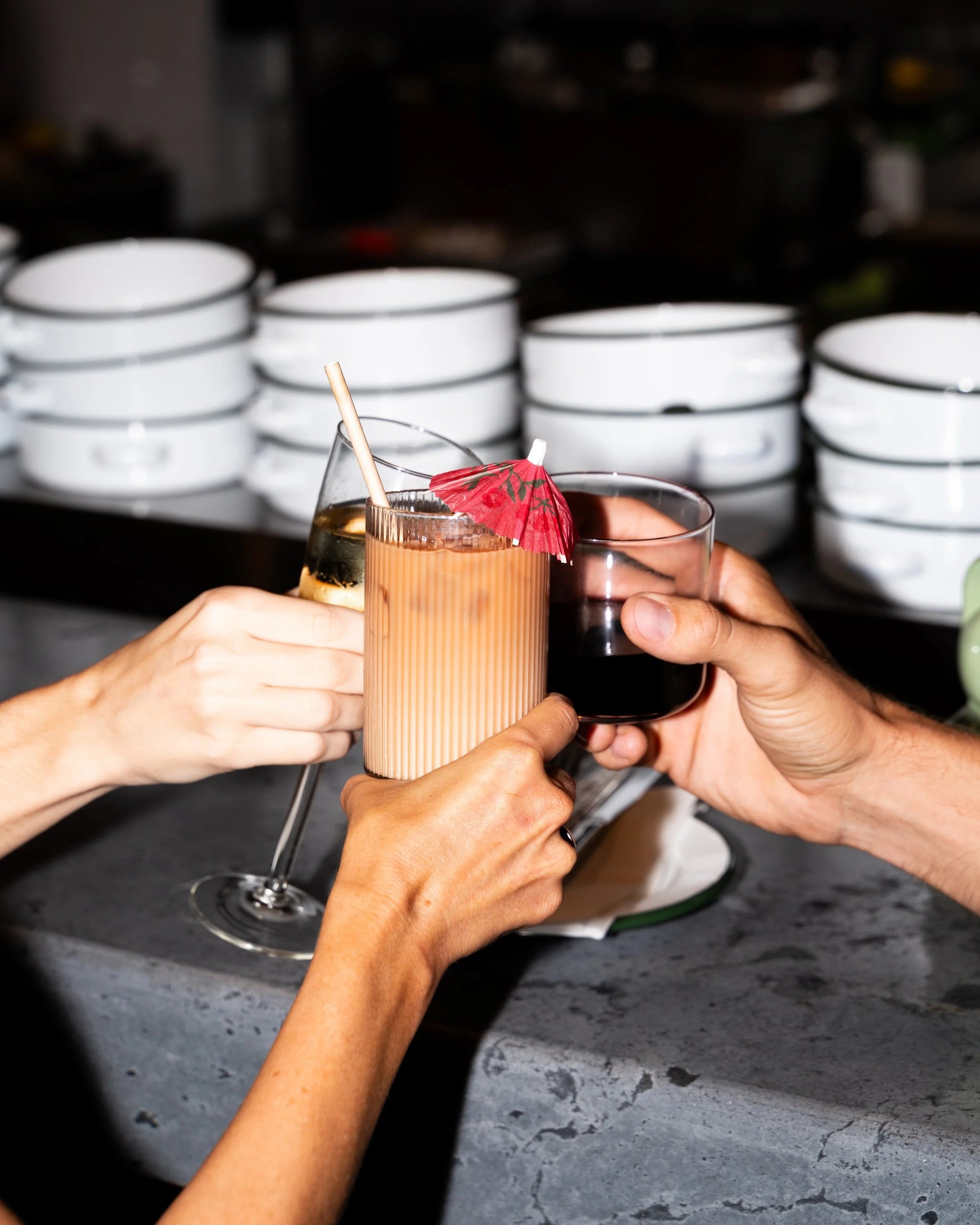 Multiple hands are holding glasses with drinks, including a cocktail with a decorative paper umbrella, in front of a stack of white dishes. The background is a dark kitchen or bar setting.