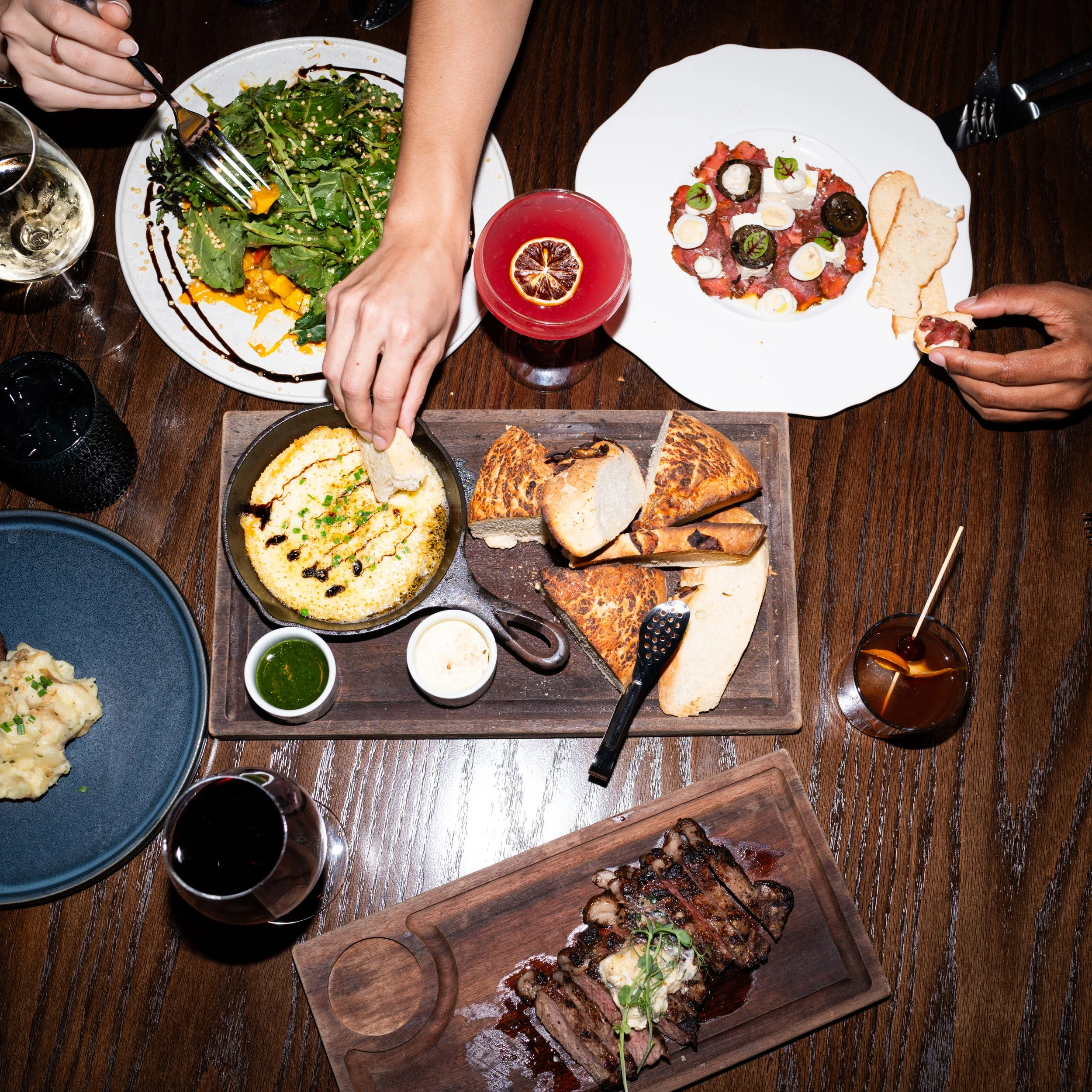 A table with various dishes including a salad, a slice of bread, a cooked dish with sauce, and drinks such as a glass of white wine and a red cocktail with a lemon slice. Food can be found at The Hew Chophouse inside the Fenway Hotel. 