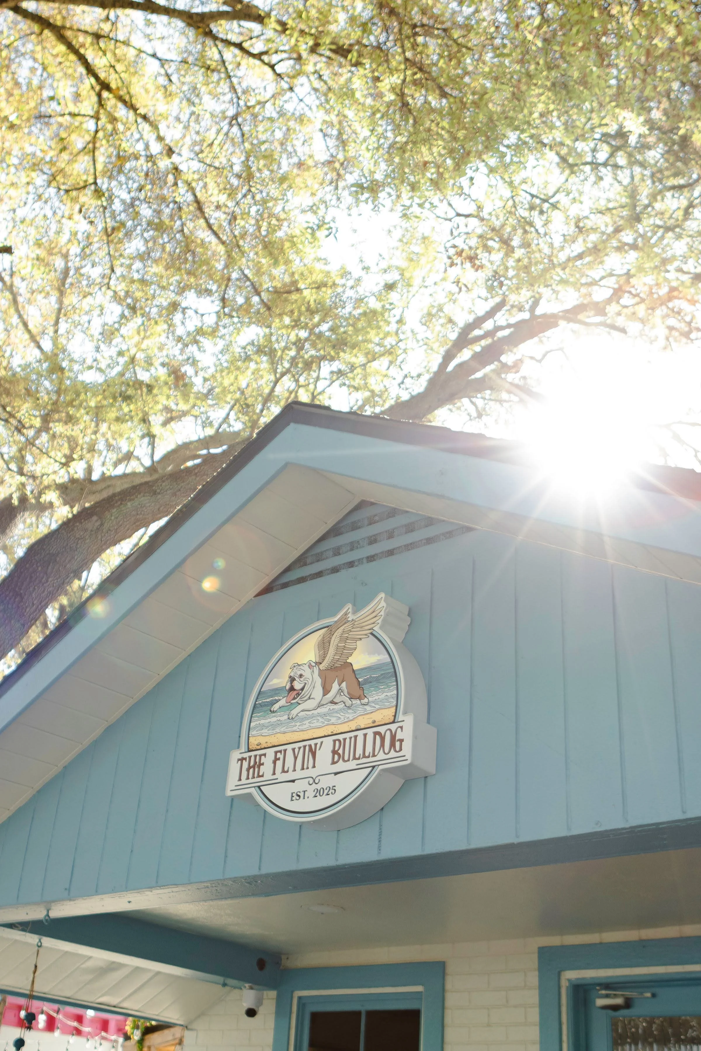Blue building with a sign featuring a bulldog logo and the text "The Flyin' Bulldog Est. 2025". Part of a tree and sunlight are visible in the background.