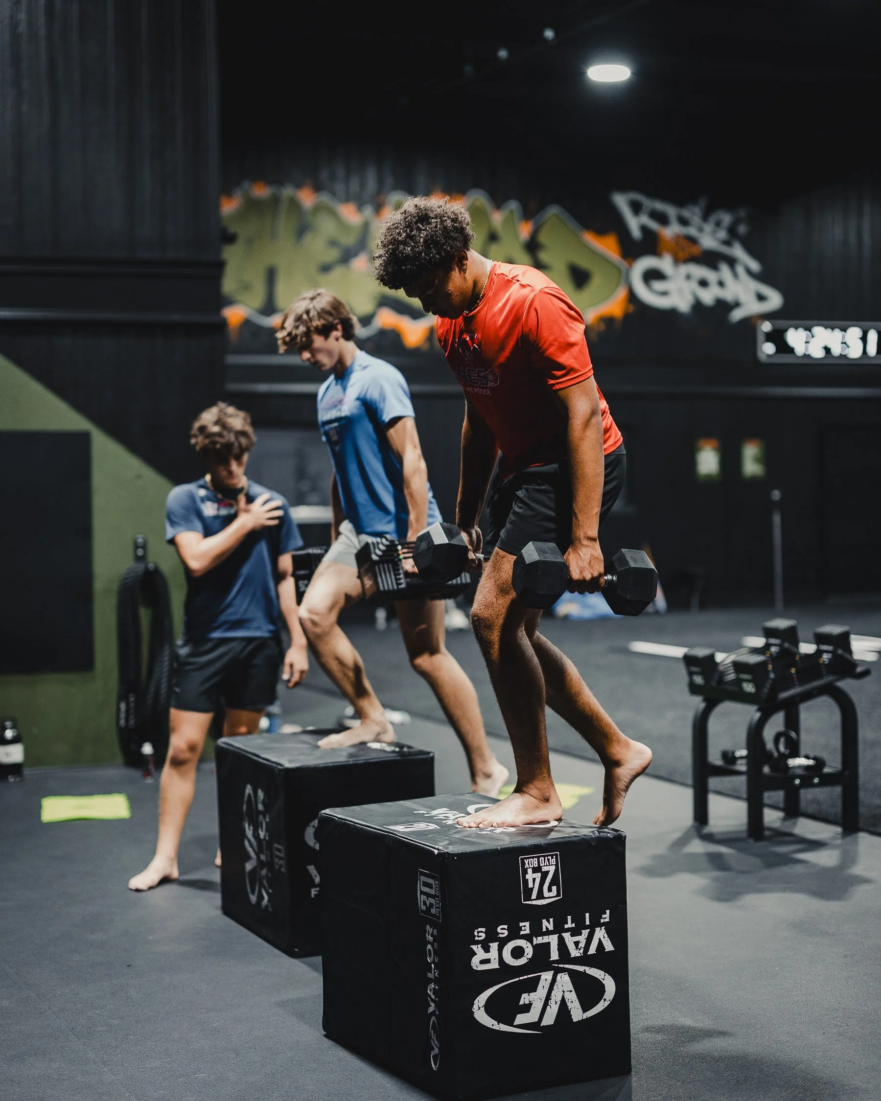 Two young men are working out at a gym. They are lifting dumbbells while standing on large padded boxes. The gym has a black interior with graffiti art on the wall that reads 'Respect & Graffiti'.