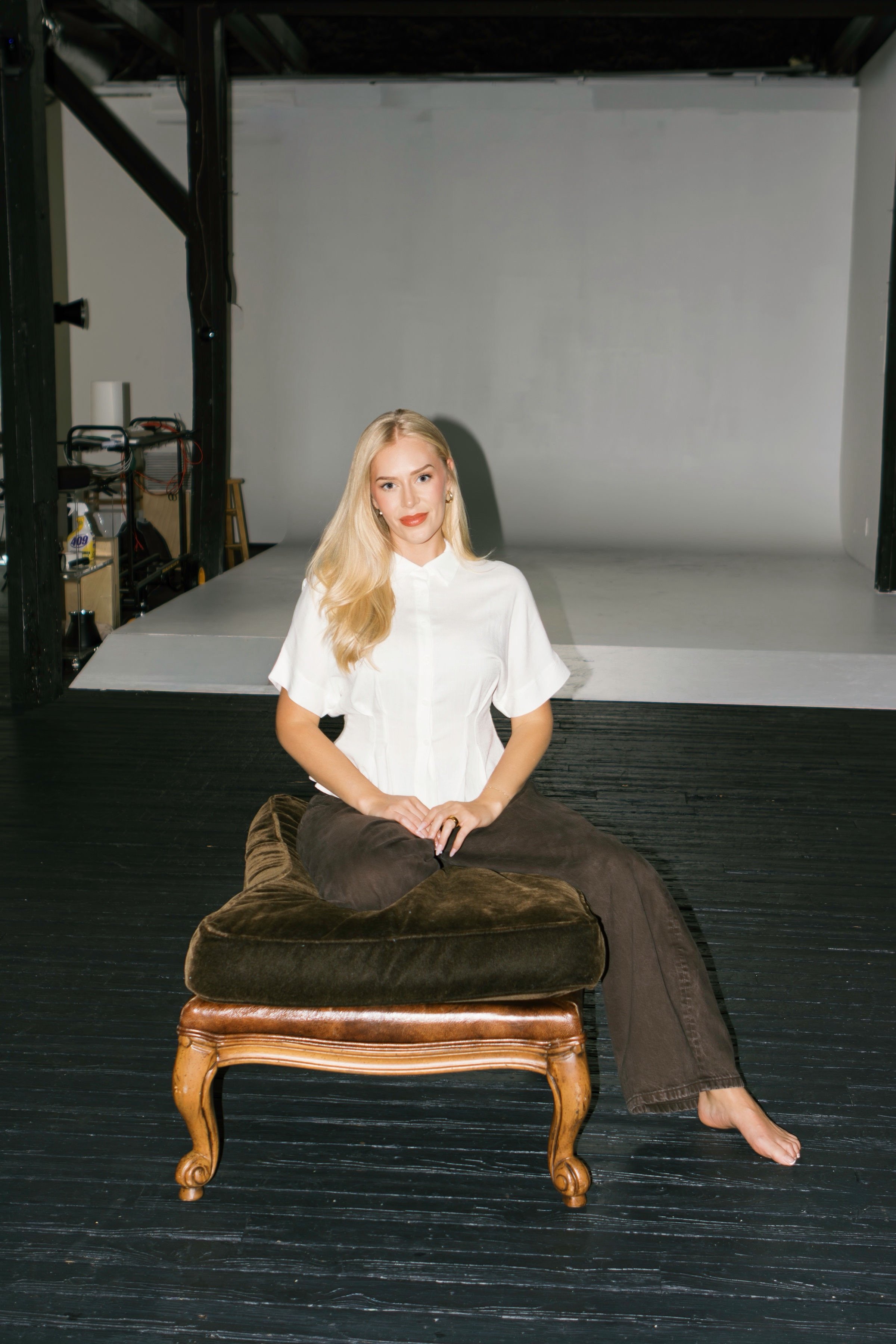 A woman with long blonde hair, wearing a white shirt and brown pants, sitting barefoot on a vintage-style wooden stool with a dark brown cushion, in front of a plain, light gray backdrop in a photography studio.