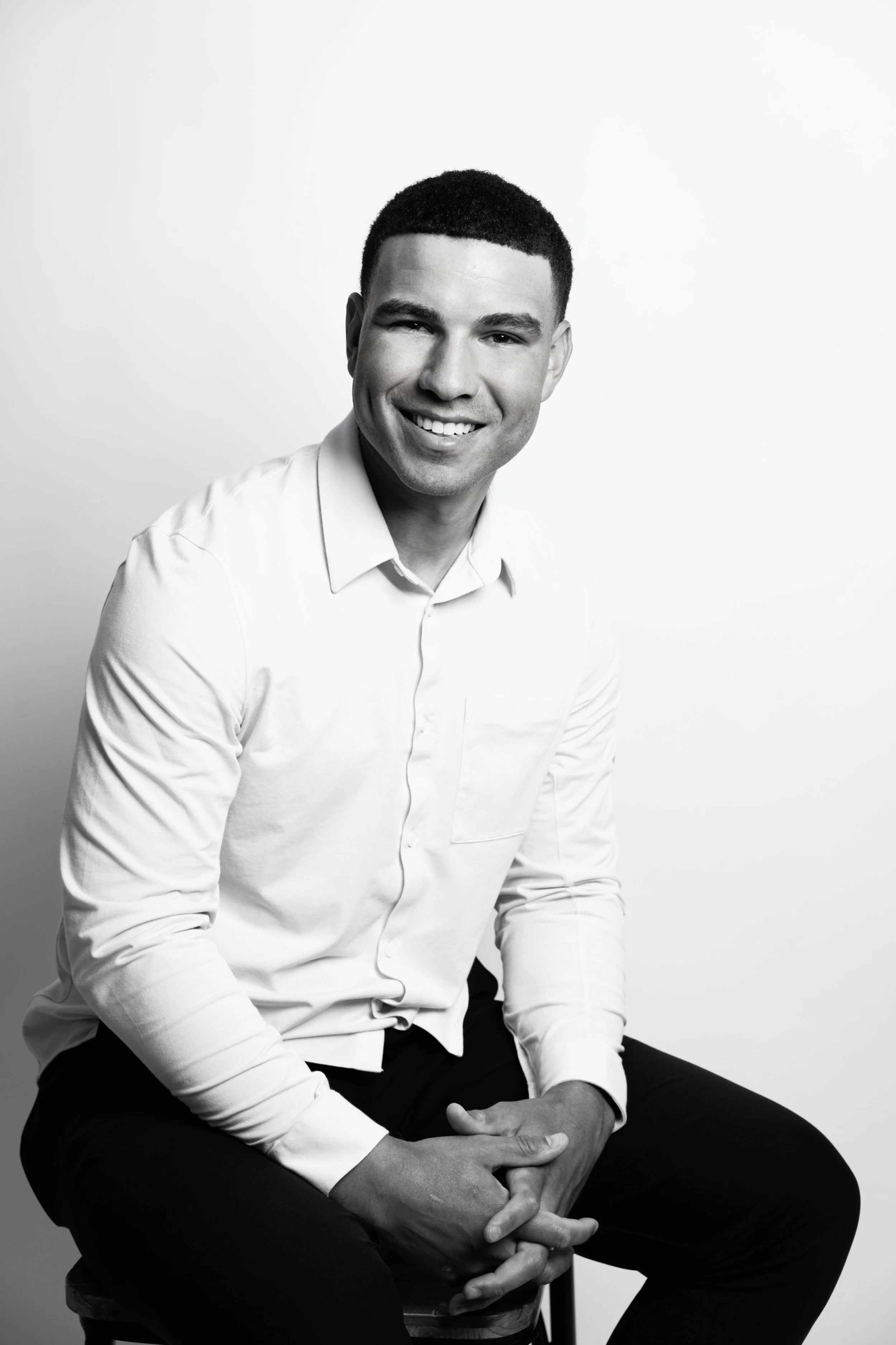 A black and white portrait of a young man with short hair, smiling, wearing a white button-up shirt and black pants. He is sitting on a stool with his hands clasped in his lap, against a plain background.