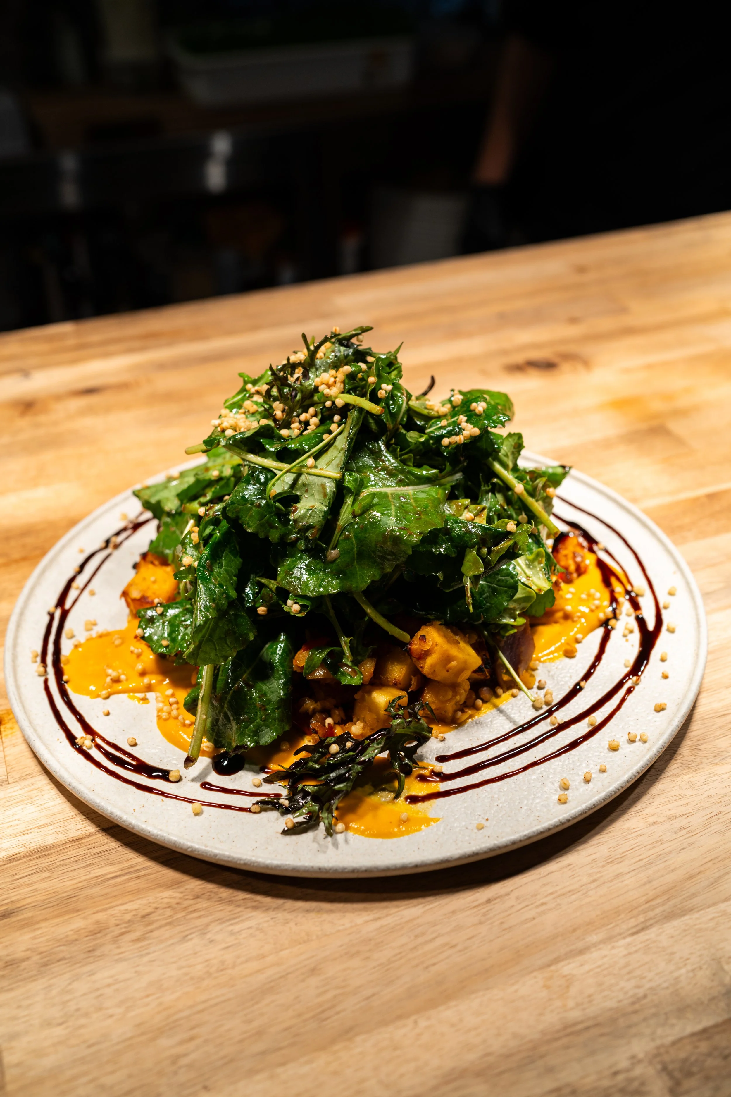A plate of food with greens topped with seeds, drizzled with sauces, on a wooden table.