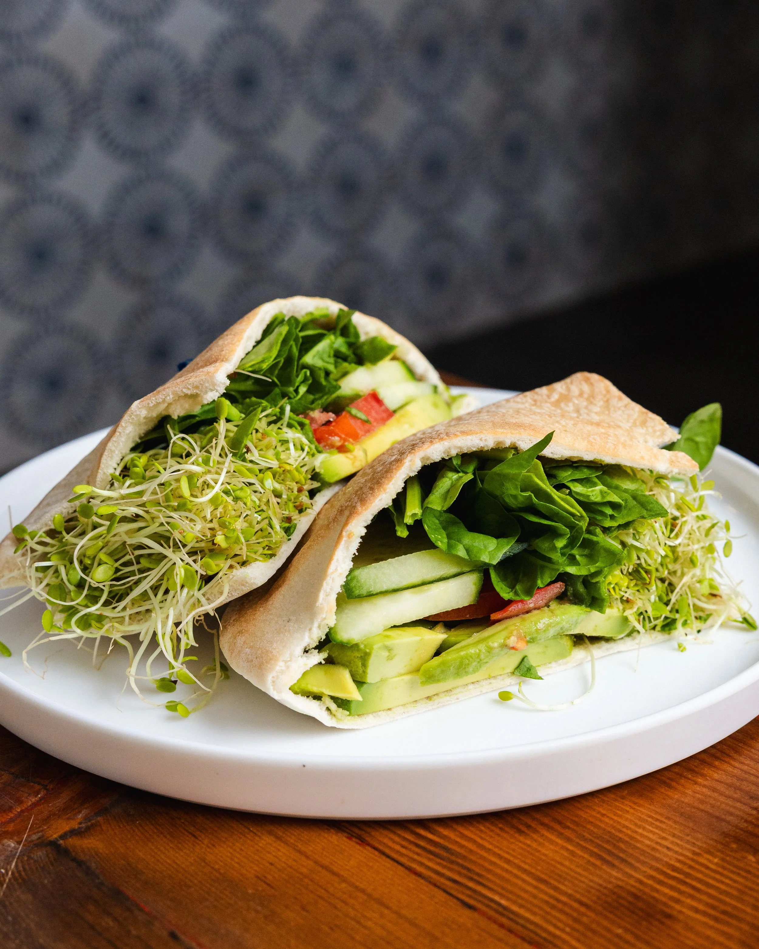 Close-up of two veggie sandwiches on a white plate with microgreens, cucumbers, tomatoes, and leafy greens, placed on a wooden table.