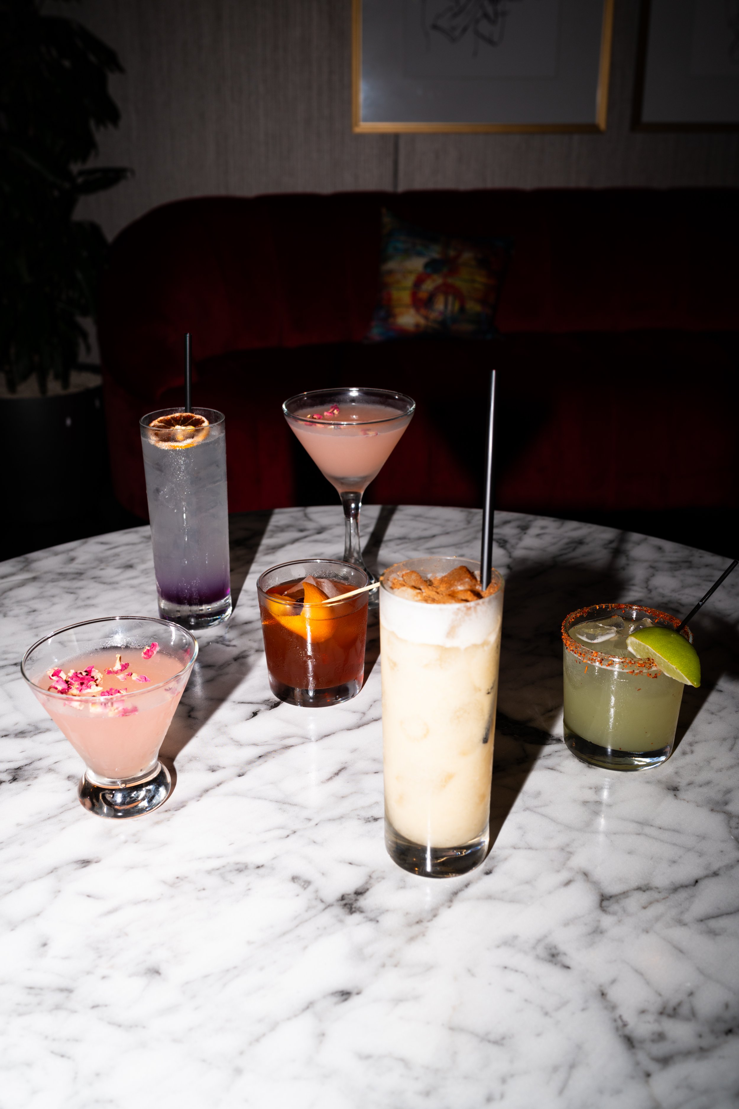 A variety of cocktails on a white marble table in a dimly lit room. The drinks include a light pink cocktail with pink flower petals, a purple and clear tall drink with a purple flower garnish, a dark amber drink with an orange twist, a tall creamy w