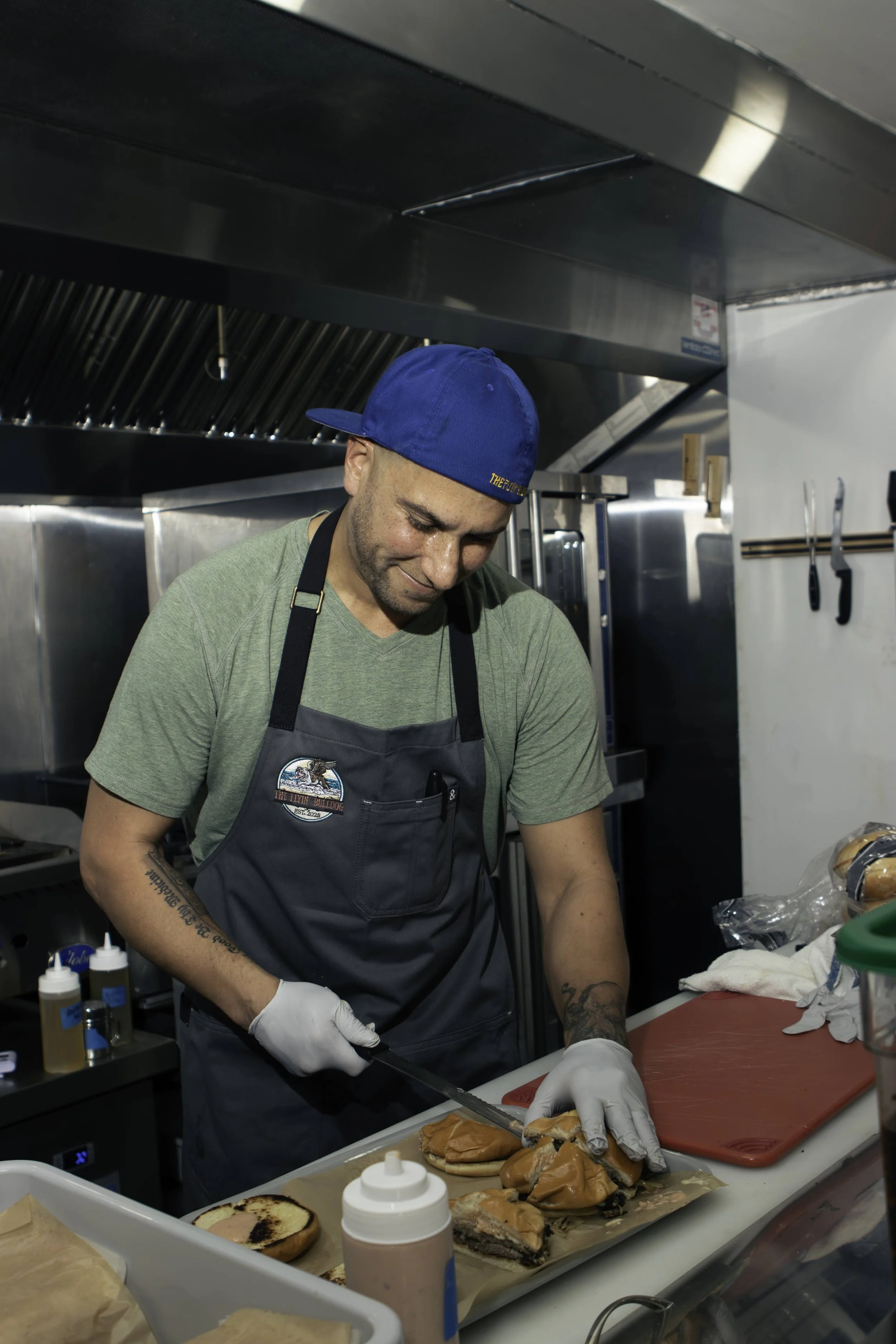 A man wearing a blue cap, green t-shirt, white gloves, and a gray apron preparing sandwiches in a commercial kitchen. The counter has ingredients, including bread and meat, with kitchen tools hanging on the wall.
