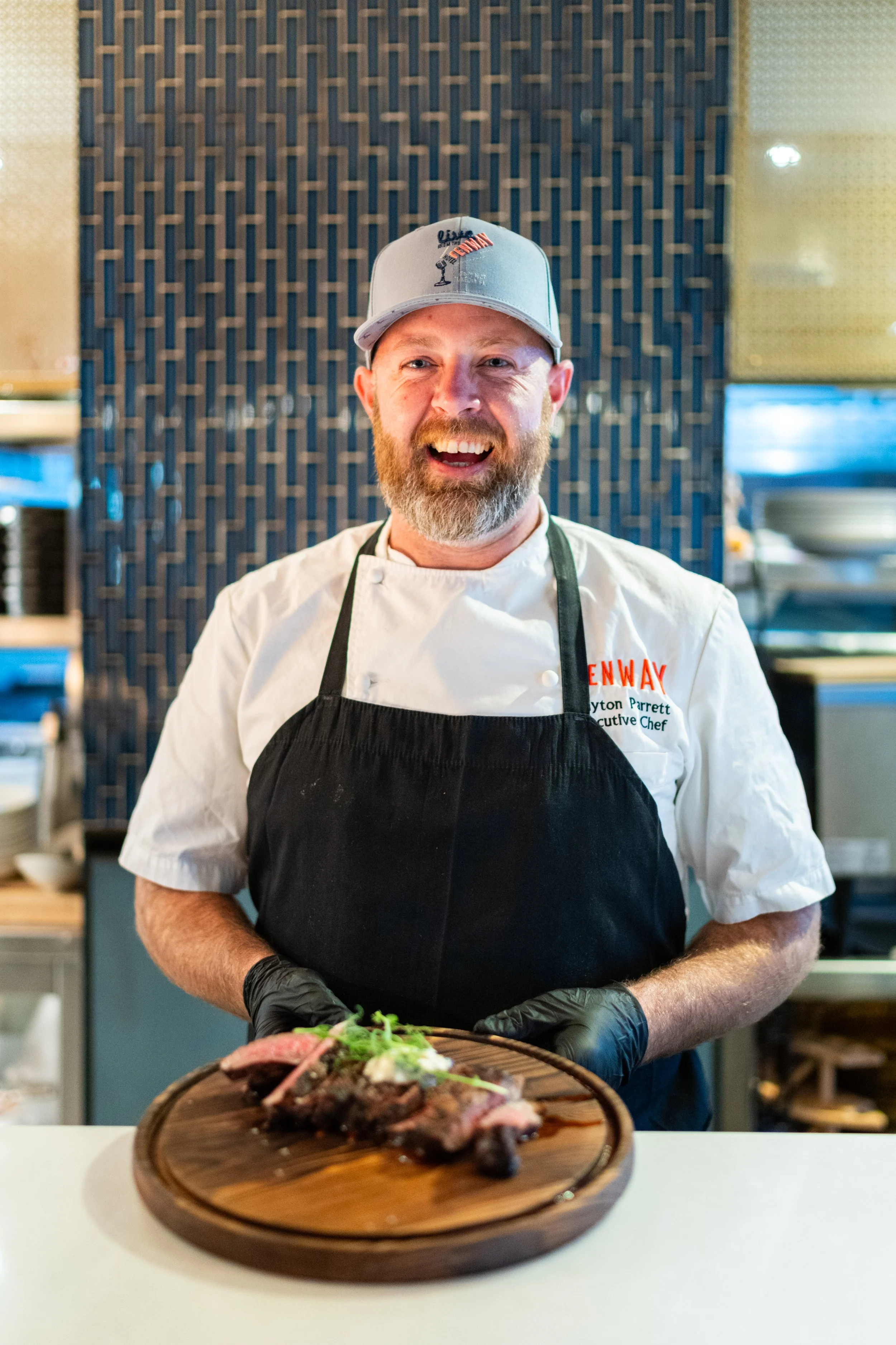 A chef wearing a gray cap, white chef coat, black apron, and black gloves, smiling while preparing steak on a wooden cutting board in a kitchen with dark tiled wall.