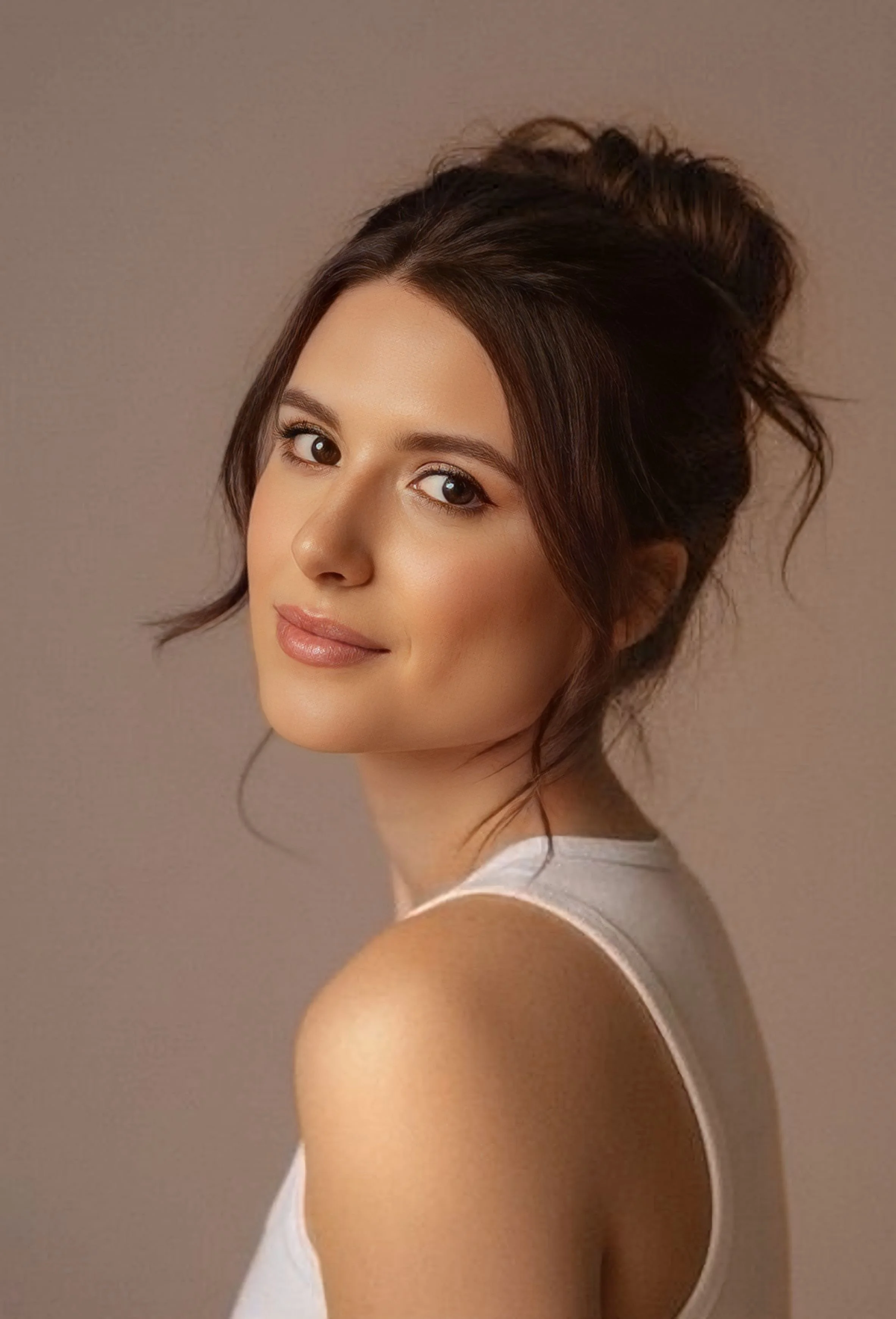 Close-up of a young woman with brown hair styled in an updo, wearing a white sleeveless top, smiling softly against a neutral background.