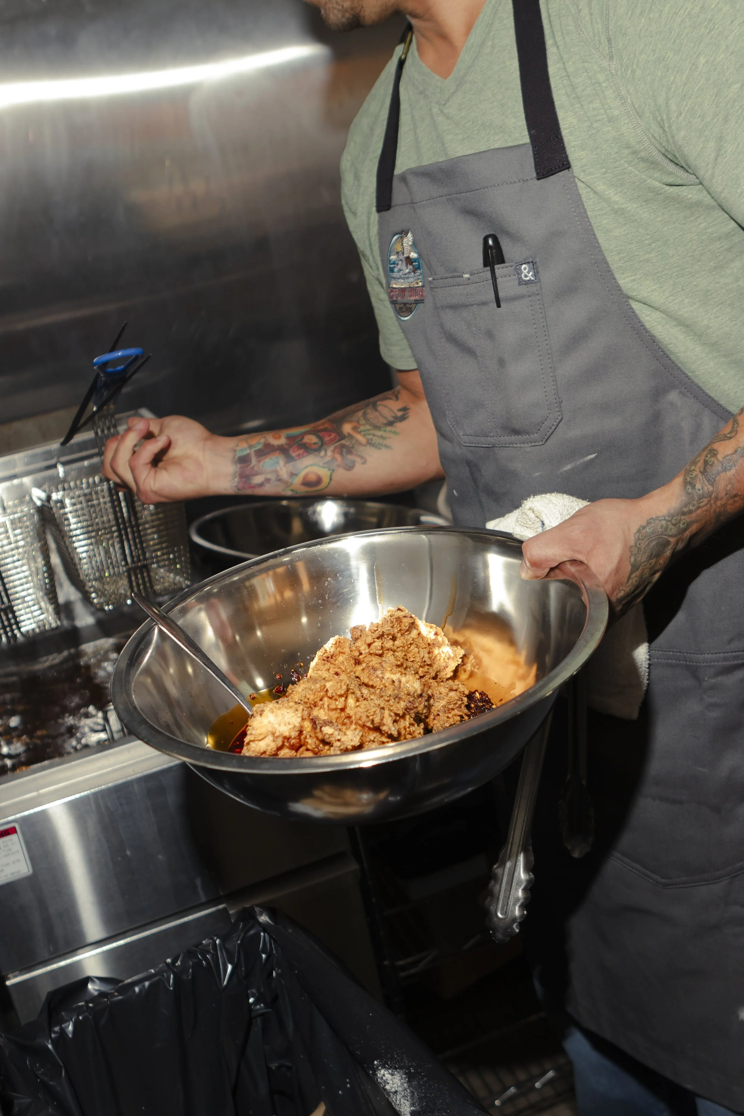 The owner of The Flyin Bulldog in Dunedin is, preparing food in a commercial kitchen. The person is holding a large stainless steel bowl with fried chicken and dipping sauce, and has a pen in a pocket on their apron.