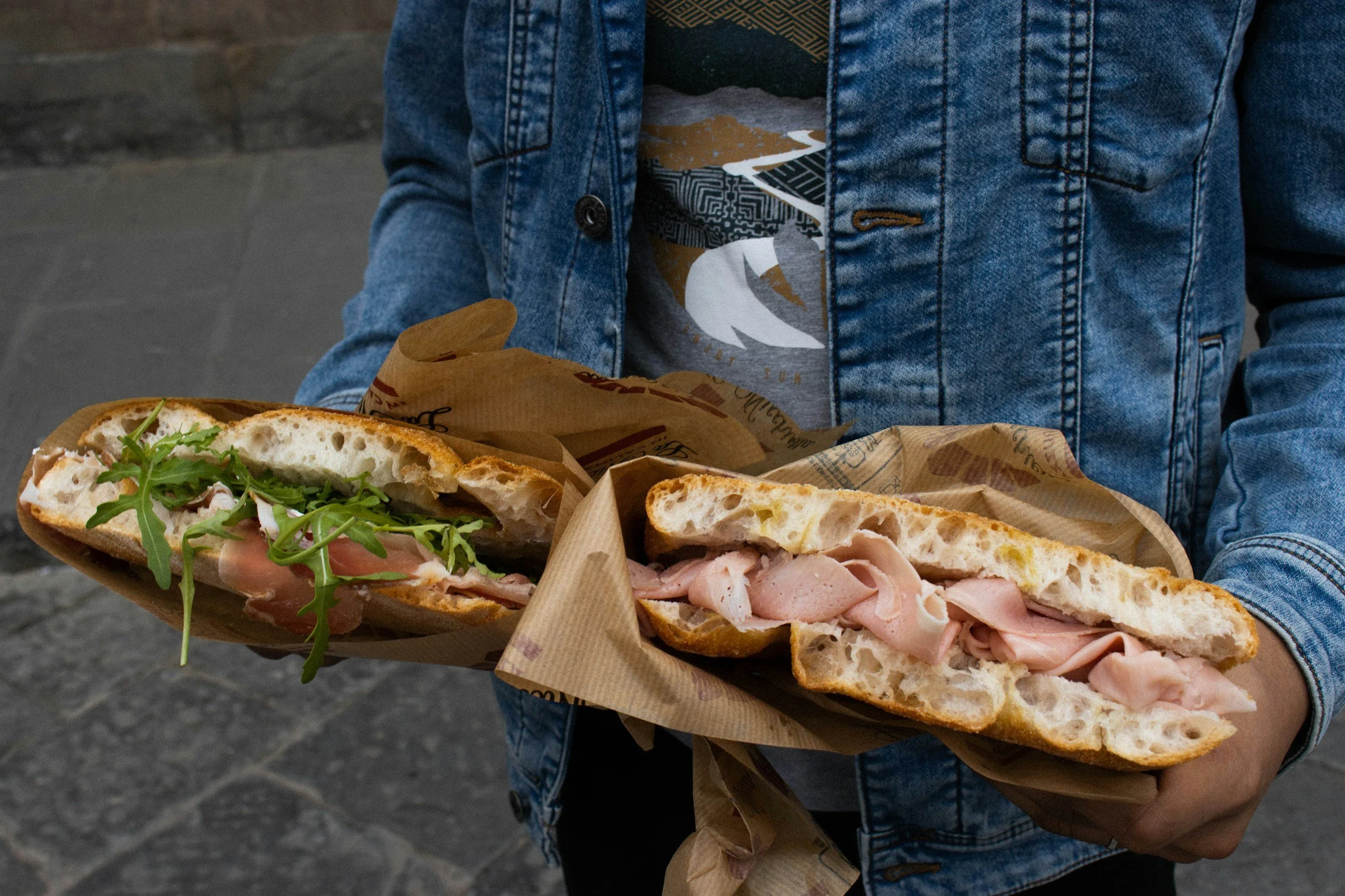 Person wearing a denim jacket holding two large sandwiches wrapped in paper, with various deli meats, arugula, and bread visible.