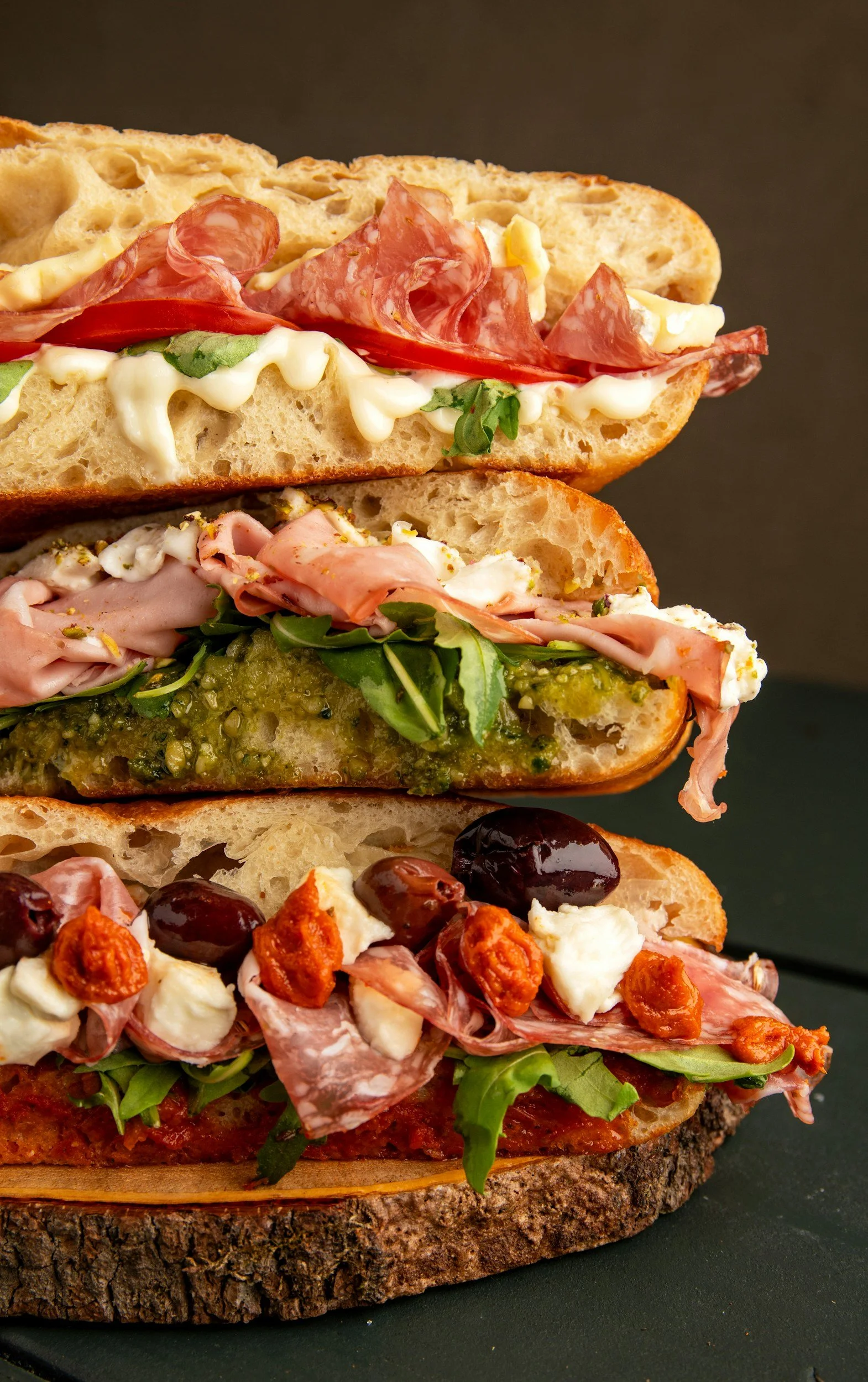 Stacked sliced artisanal bread sandwiches with various fillings, including meats, cheeses, vegetables, and toppings on a dark surface.