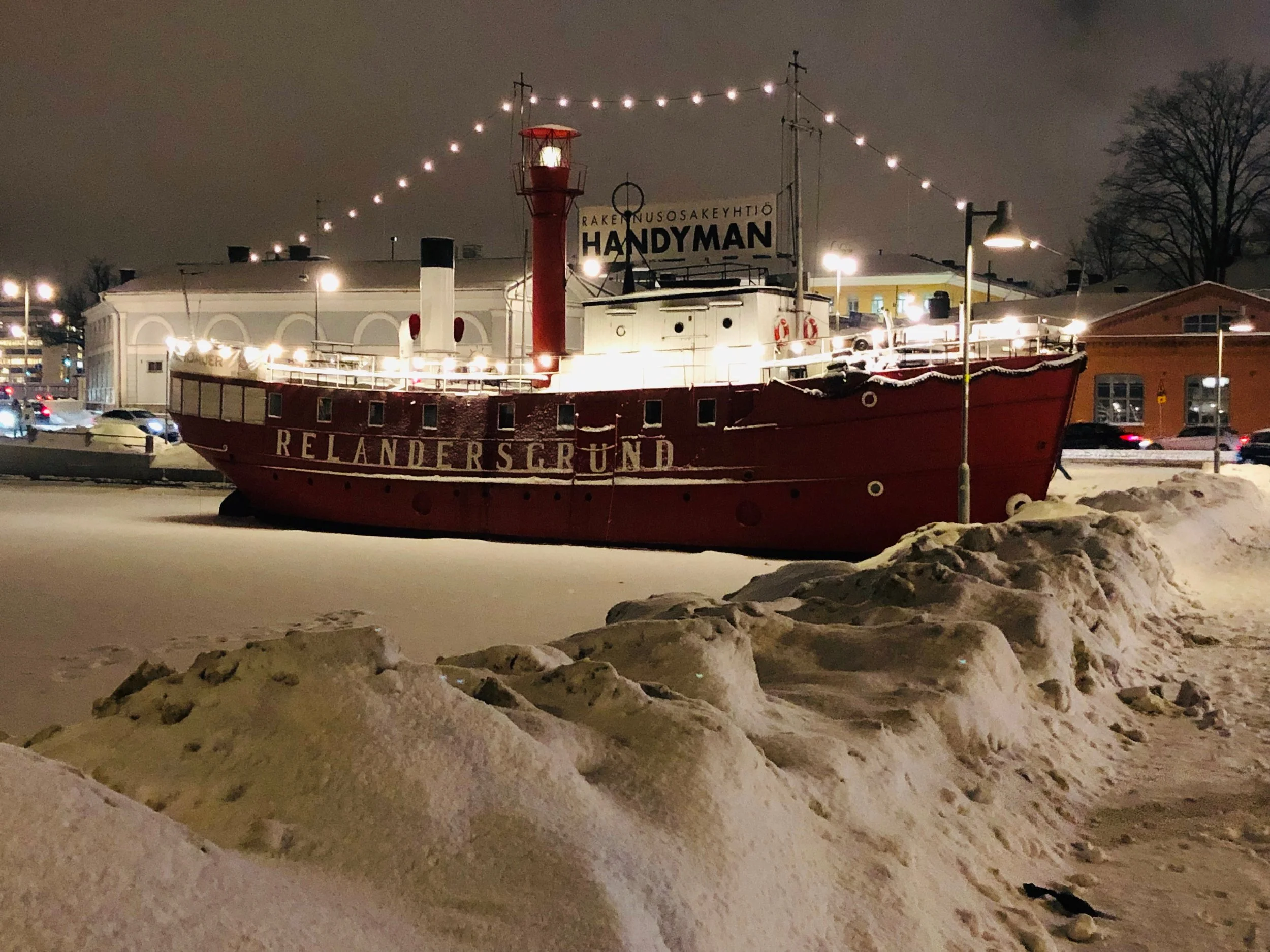 A boat with the name 'Relandersgrund' on the side, docked near snow-covered ground at night, illuminated by string lights and nearby streetlights, with buildings and trees in the background.