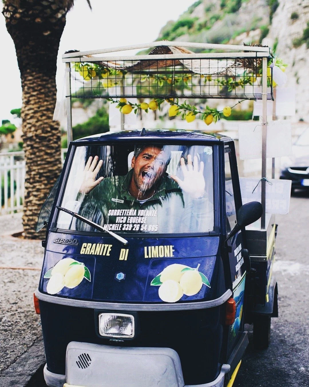 A small three-wheeled vehicle, possibly an auto rickshaw, decorated with lemon graphics and text in Italian, with a man inside waving and making a funny face. The vehicle is parked outdoors near a tree with a lemon garland and a mountain in the background.