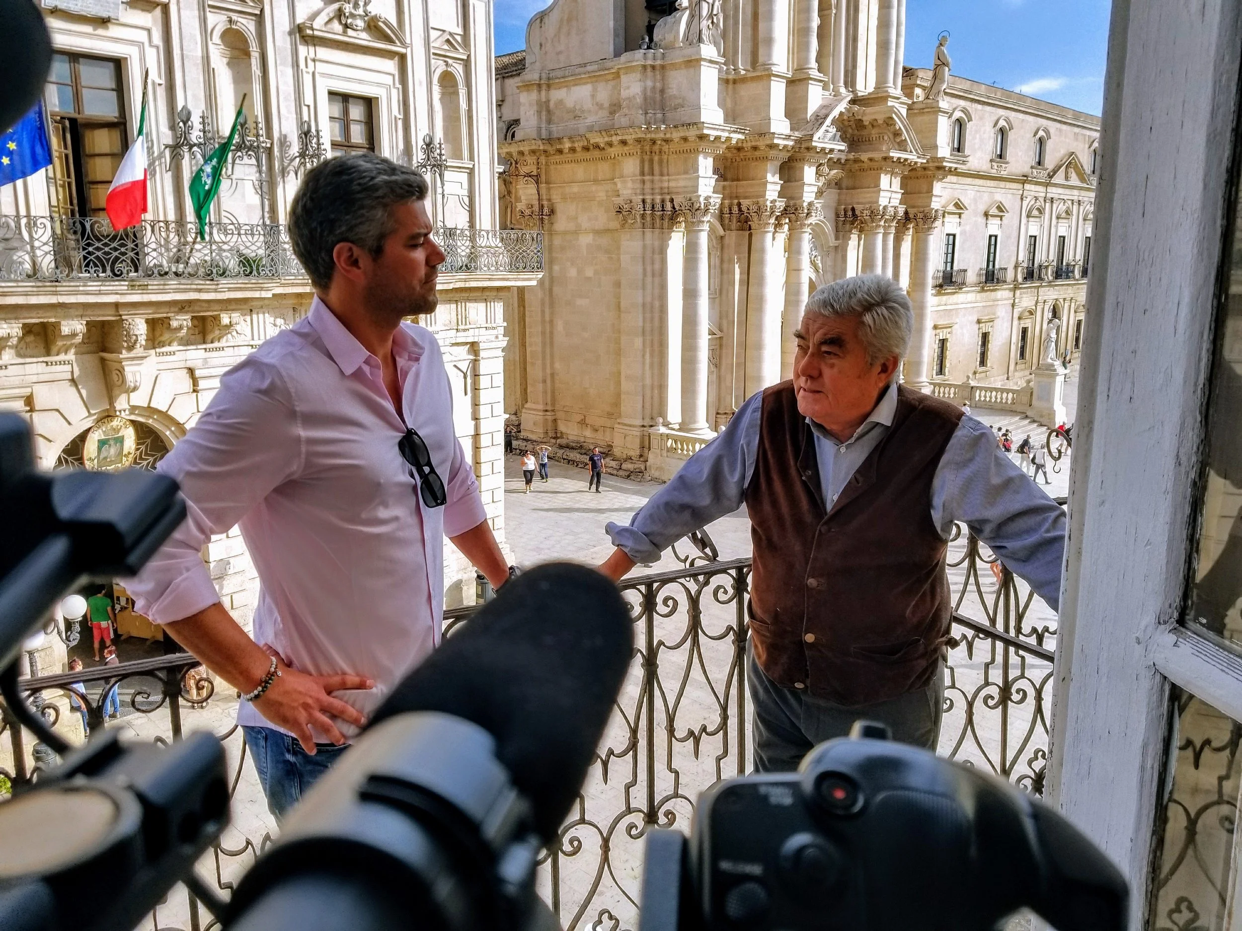 Two men are standing on a balcony overlooking historic European architecture with statues, columns, and flags in the background. One man has gray hair and is wearing a vest, while the other has dark hair and is wearing a pink shirt. The photo appears to be taken from behind a camera or recording device.
