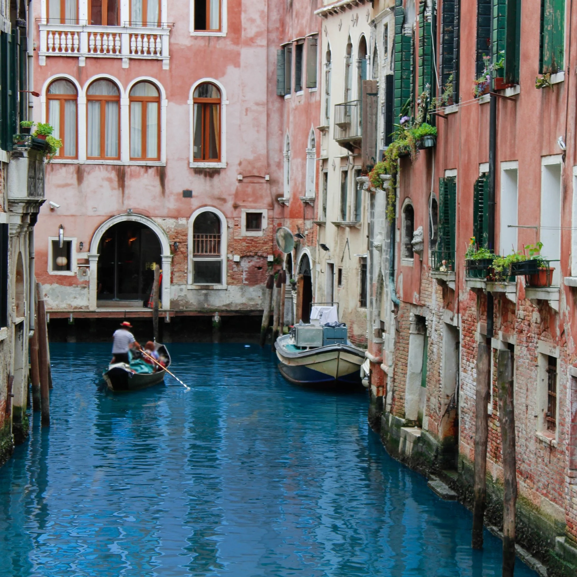 A narrow canal in Venice, Italy, with colorful old buildings on both sides, boats on the water, and a person in a boat in the foreground.