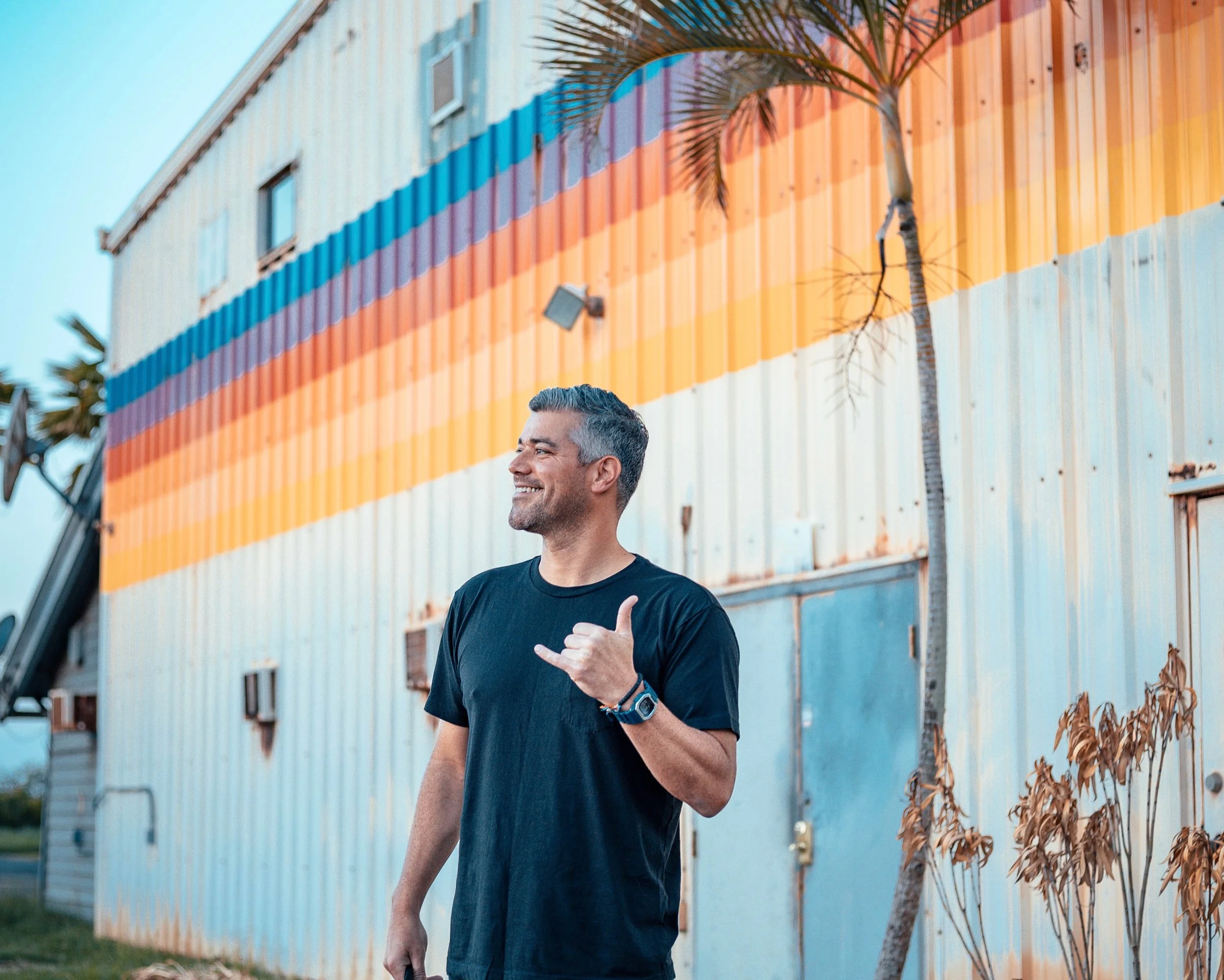 A man with gray hair smiling and making a hand gesture, standing outdoors in front of a colorful striped building with a palm tree nearby.