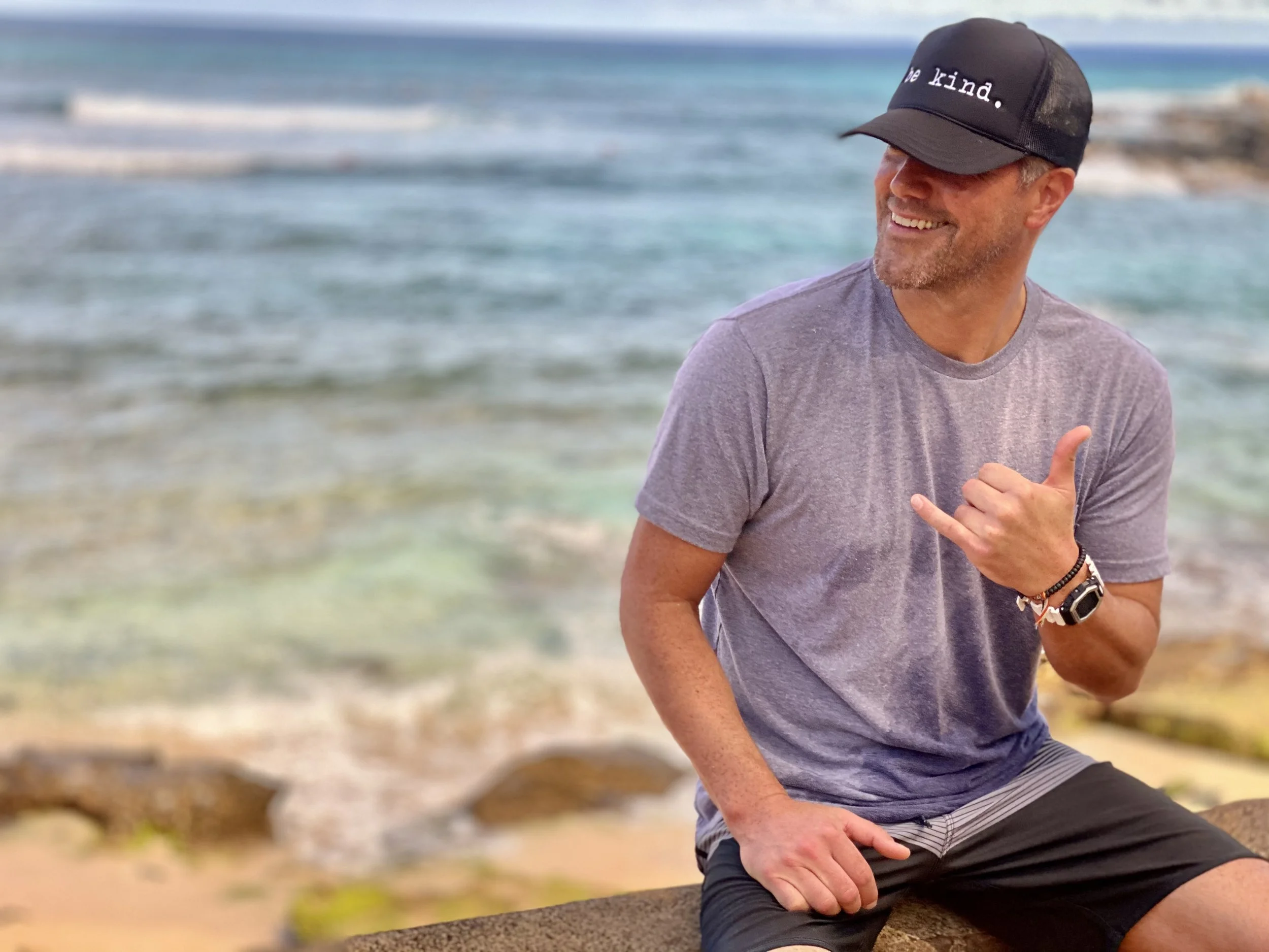 A smiling man wearing a black cap with the words 'be kind.' and a gray T-shirt, sitting by the beach with waves in the background.