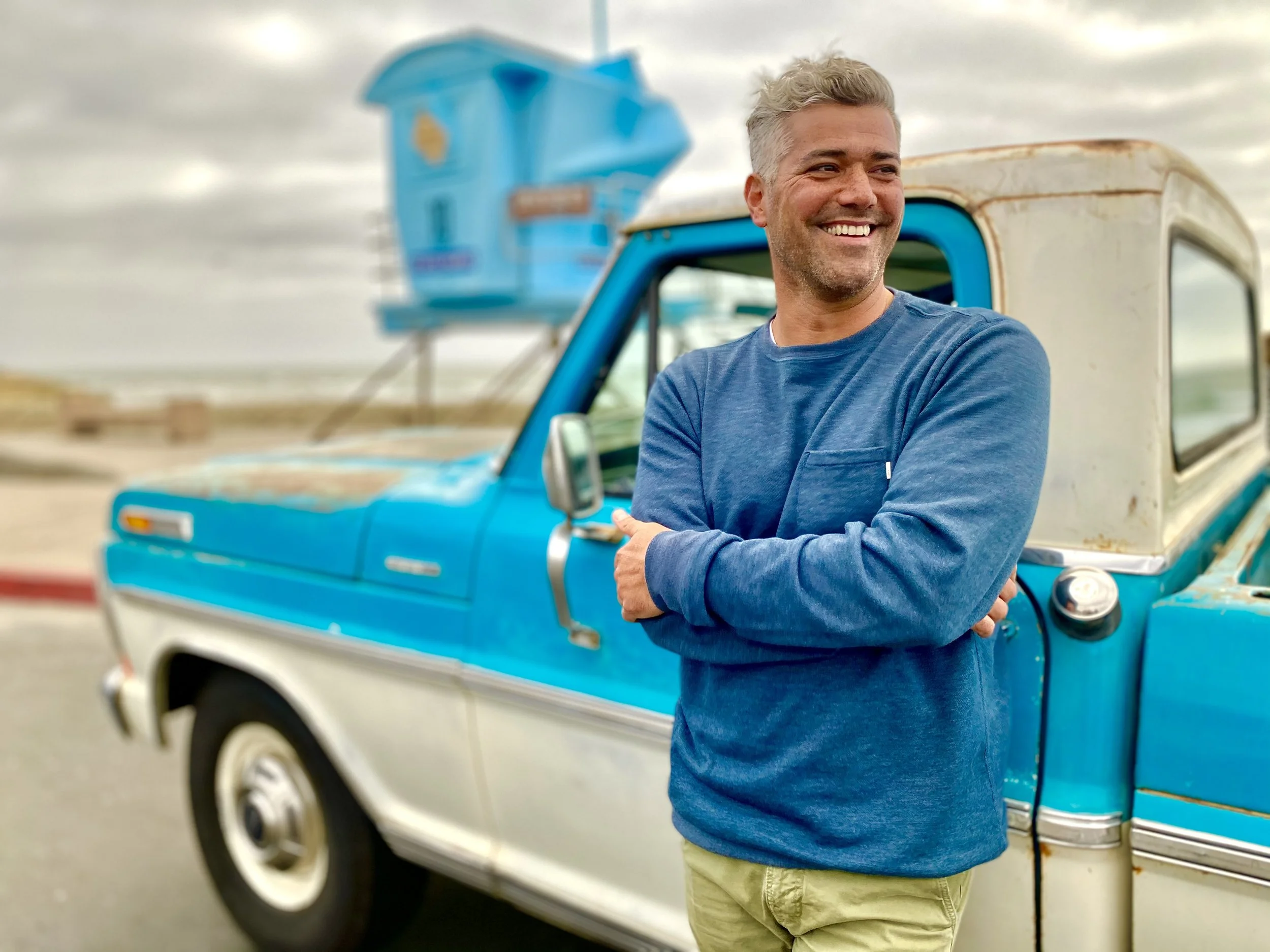 A man with gray hair and a beard smiling and crossing his arms, standing in front of a vintage blue and white pickup truck outdoors.