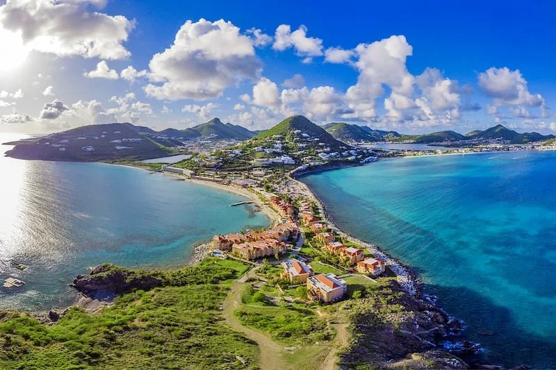 Aerial view of a coastal island with colorful houses along the shoreline, lush green hills, and blue ocean waters under a partly cloudy sky.