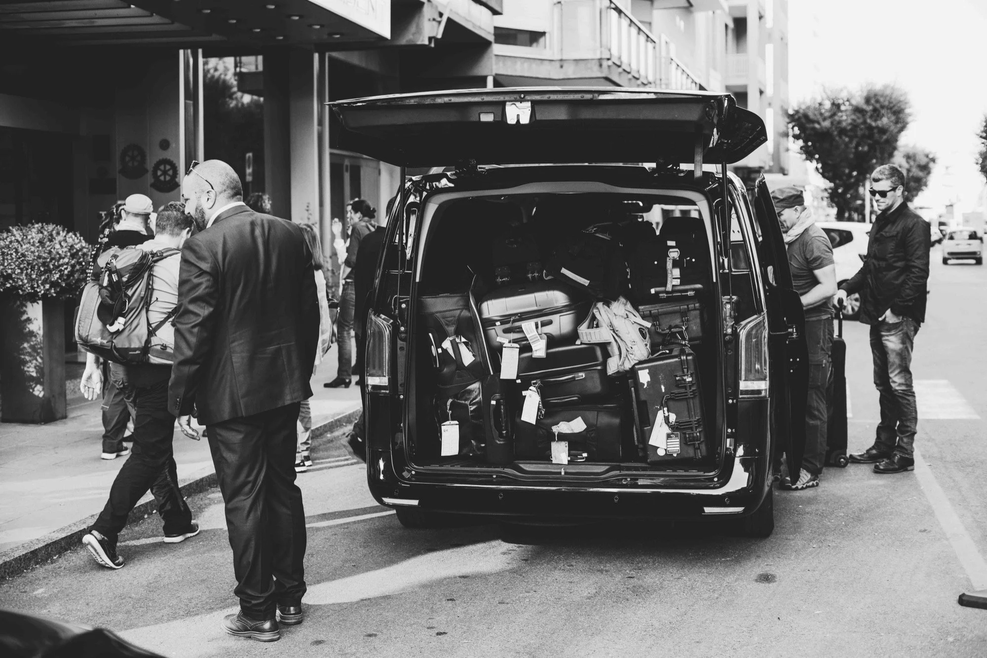 A black and white photo of people loading or unloading equipment from a van on a city street. The van's back doors are open, revealing various gear inside. Several people are gathered around, some looking into the van and others walking nearby.