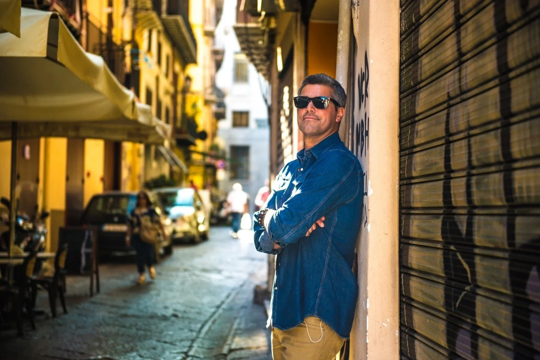 Paulino Duran wearing sunglasses, a denim jacket, and khaki pants leaning against a graffiti-covered wall on a narrow city street, with outdoor seating and parked cars in the background.