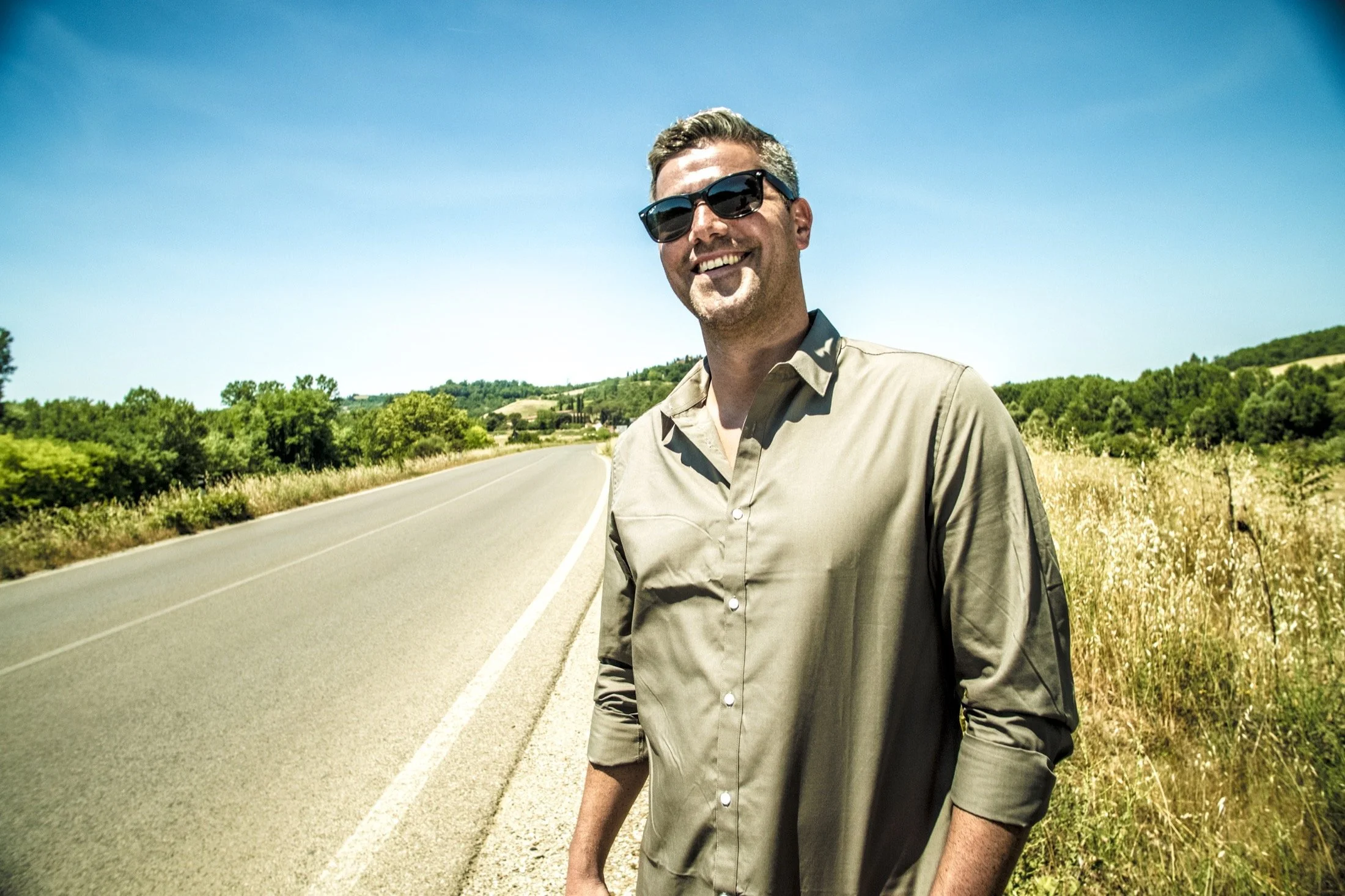 Paulino Duran wearing sunglasses and a beige shirt standing on the side of a rural road with at least four lanes, surrounded by green trees and hills under a clear blue sky.