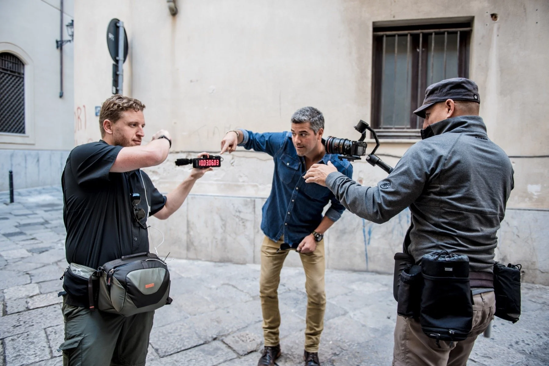 Three men with filming equipment standing on a cobblestone street against a beige wall, one man holding a digital device, another pointing at it, and the third operating a camera.