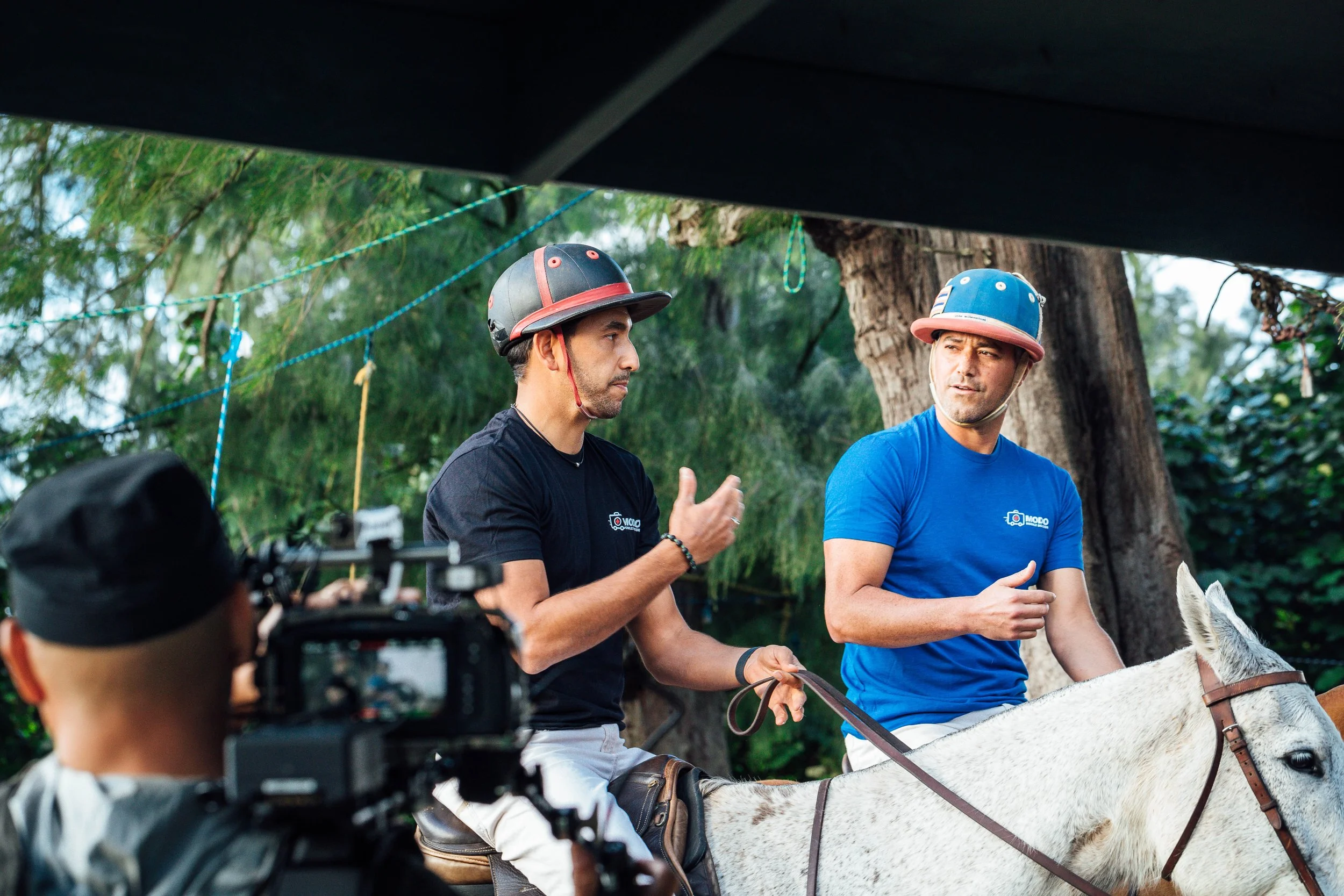 Two men wearing helmets and casual clothes sit on a white horse, engaging in a conversation outdoors with trees in the background, while a camera captures the scene.
