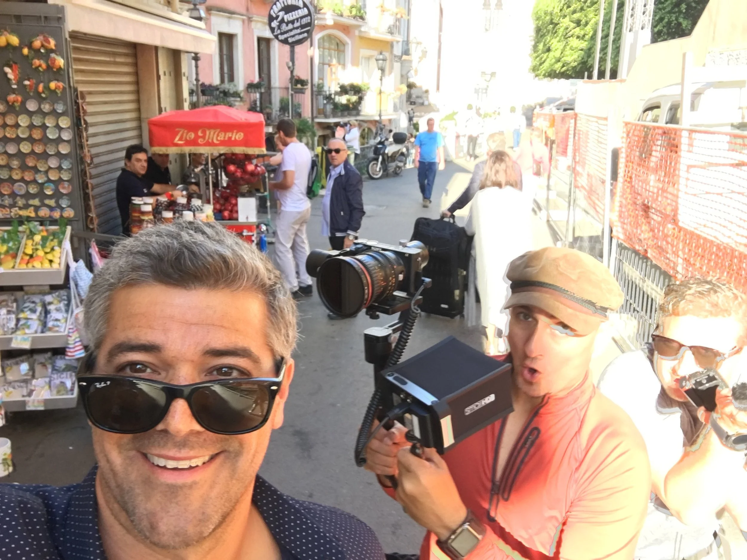 Selfie of two men in a crowded city street, one wearing sunglasses and the other holding a camera mounted on a handheld device. The background shows a street side vendor with a red umbrella, buildings, and pedestrians.