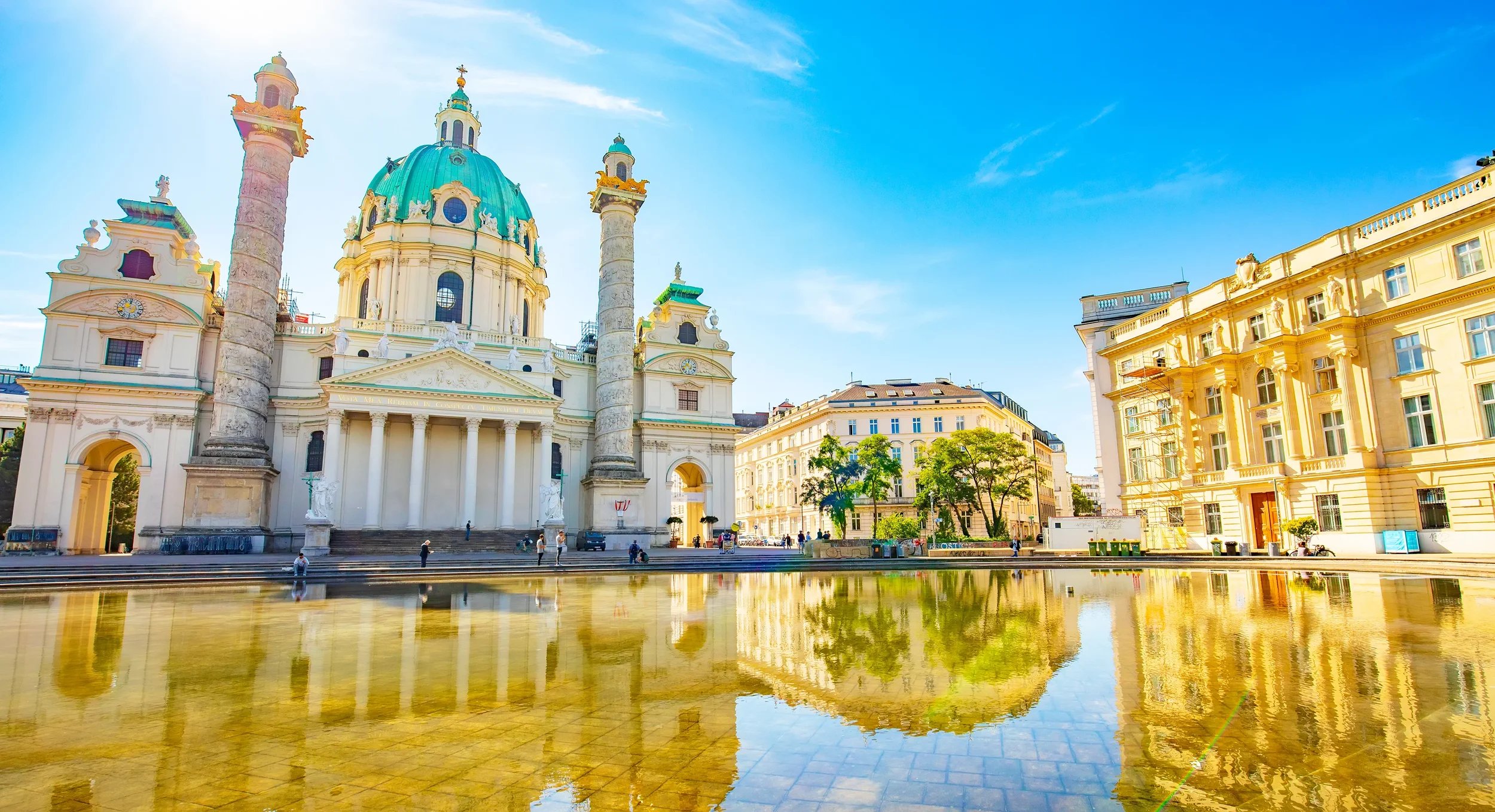 Baroque-style church with a blue-green dome and two tall columns in a city square, surrounded by yellow and cream buildings, with a reflecting water feature in the foreground and a clear blue sky.