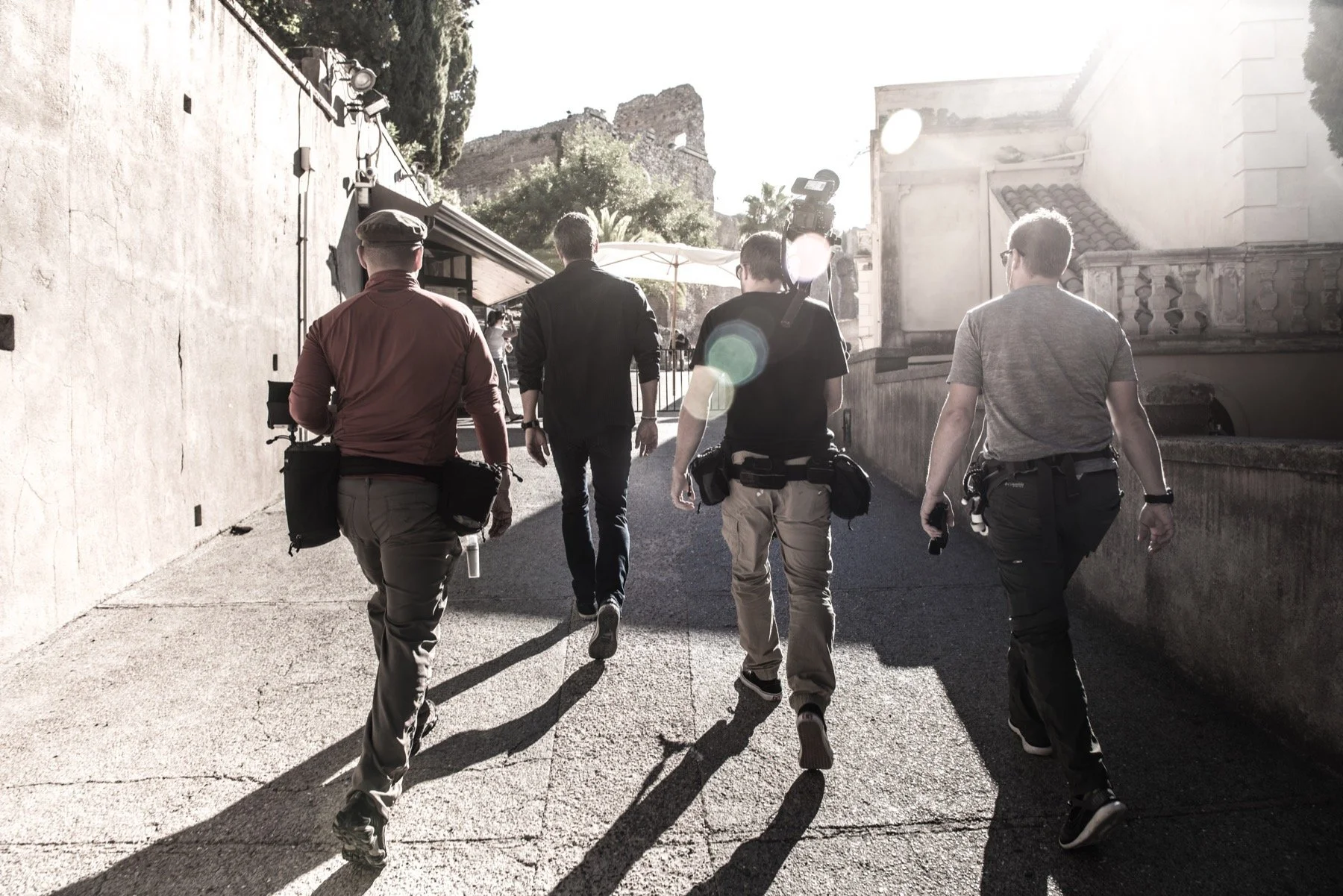 Five men walking down a sunlit alley, with some carrying cameras and equipment, suggesting they are part of a film crew or production team.