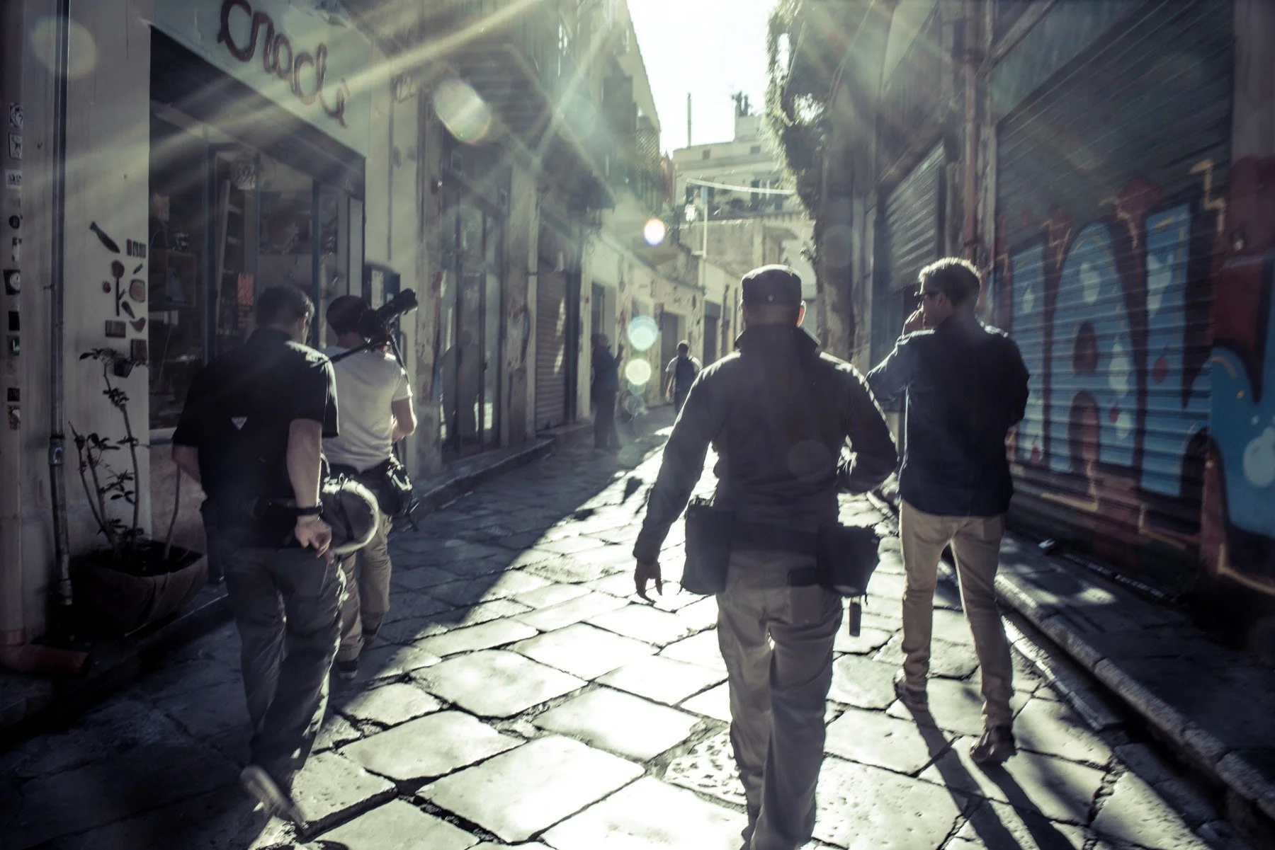 A group of men walking down a narrow cobblestone street in an urban area, with sunlight streaming through and graffiti on storefronts.