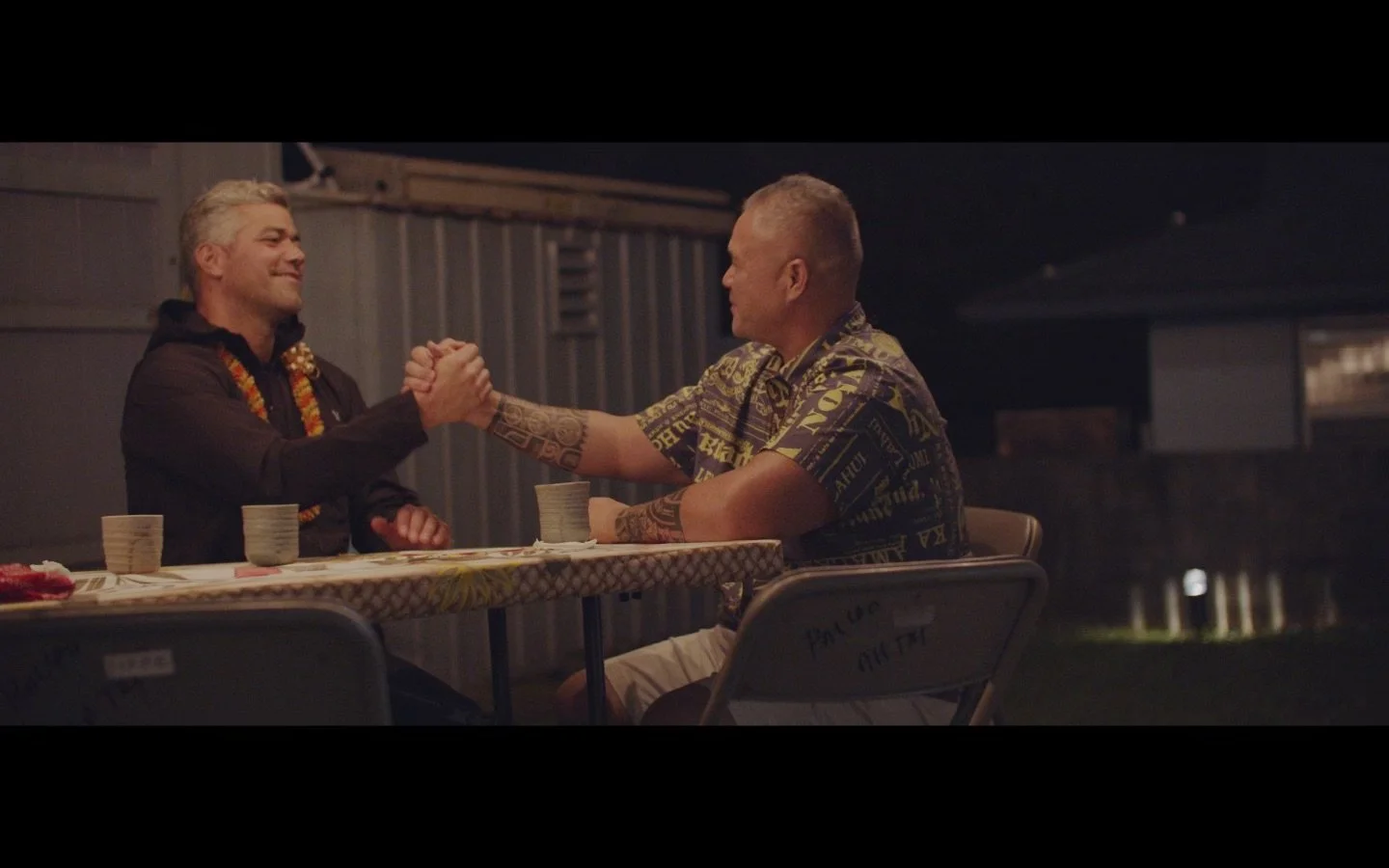 Two men sitting at a table outdoors at night, smiling and shaking hands. They are wearing leis around their necks, with drinks and food on the table.