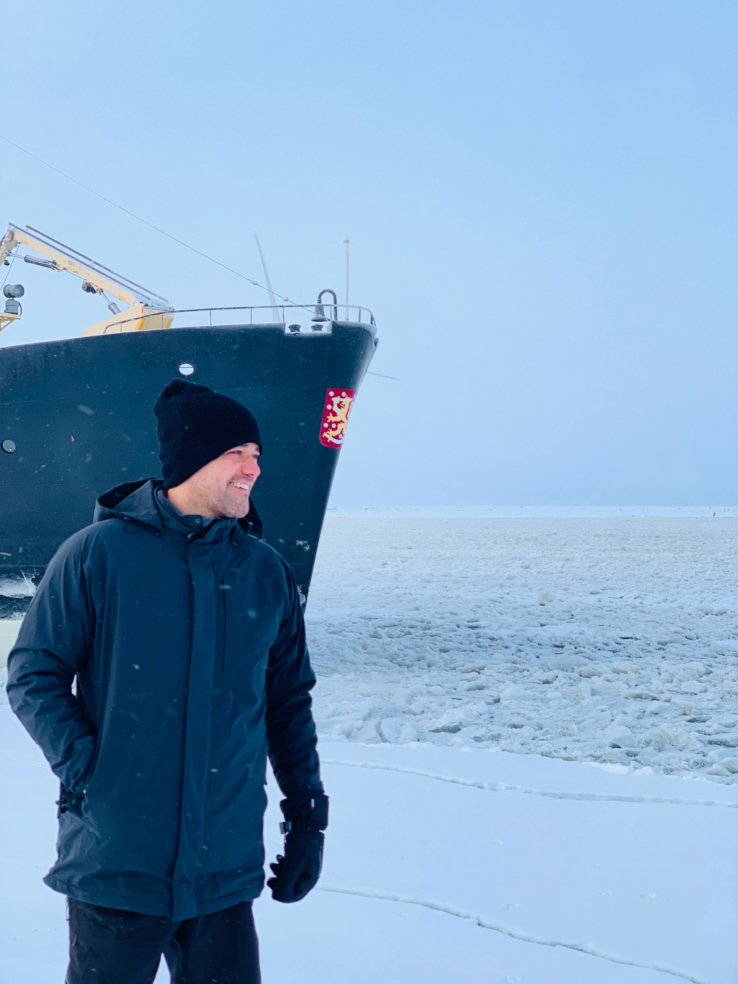 A man wearing a black winter jacket, black beanie, and gloves standing on snow-covered ice with a large ship in the background on a cloudy day.