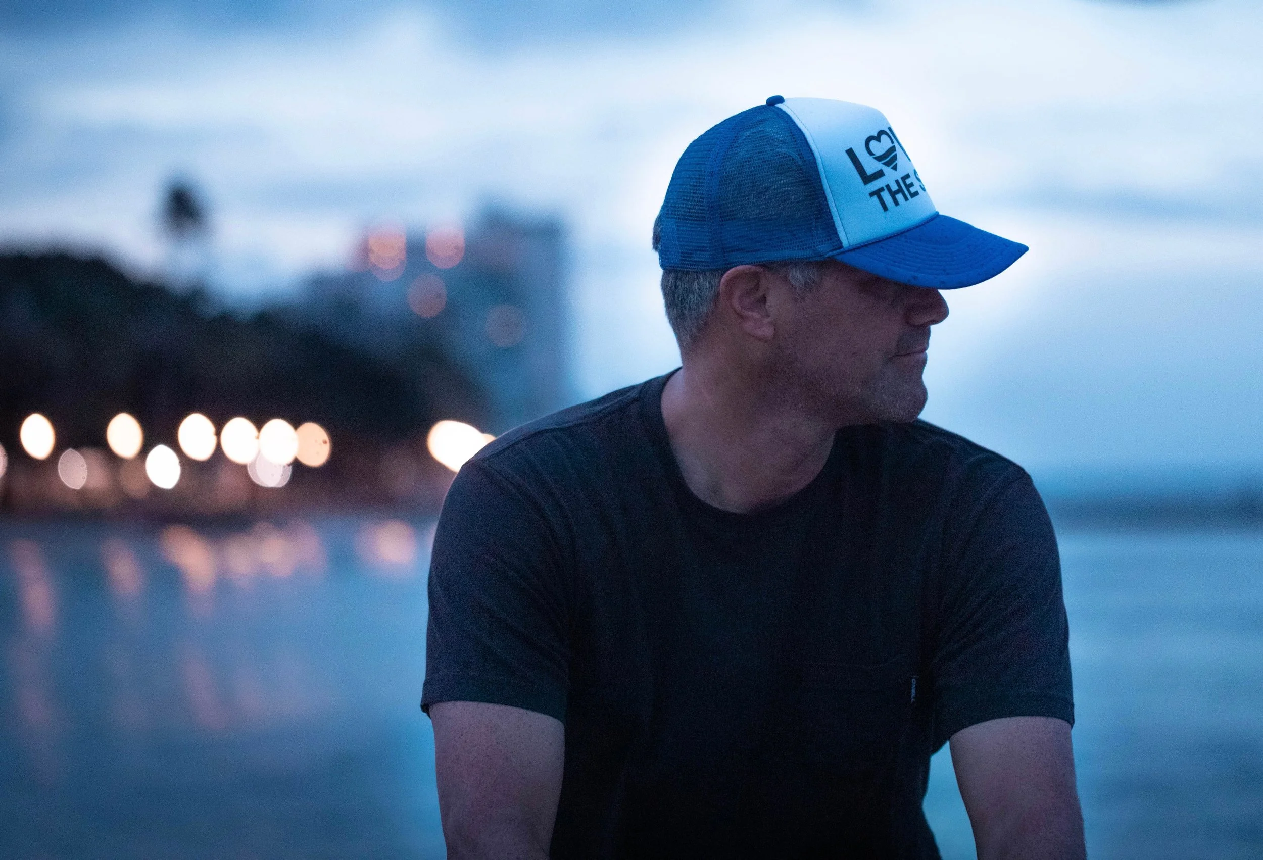 A man wearing a blue trucker hat and a black t-shirt, looking to the right, with a blurred background of city lights at dusk.