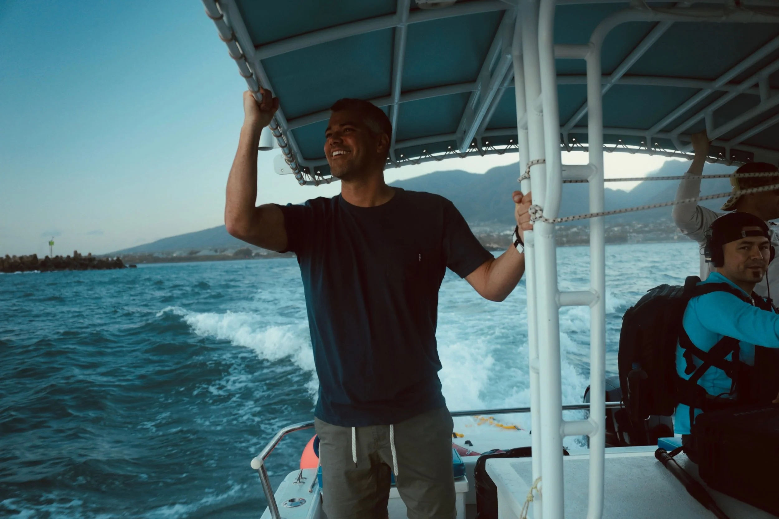 A man smiling and holding onto a boat's roof frame while sailing on the water with mountains in the background.