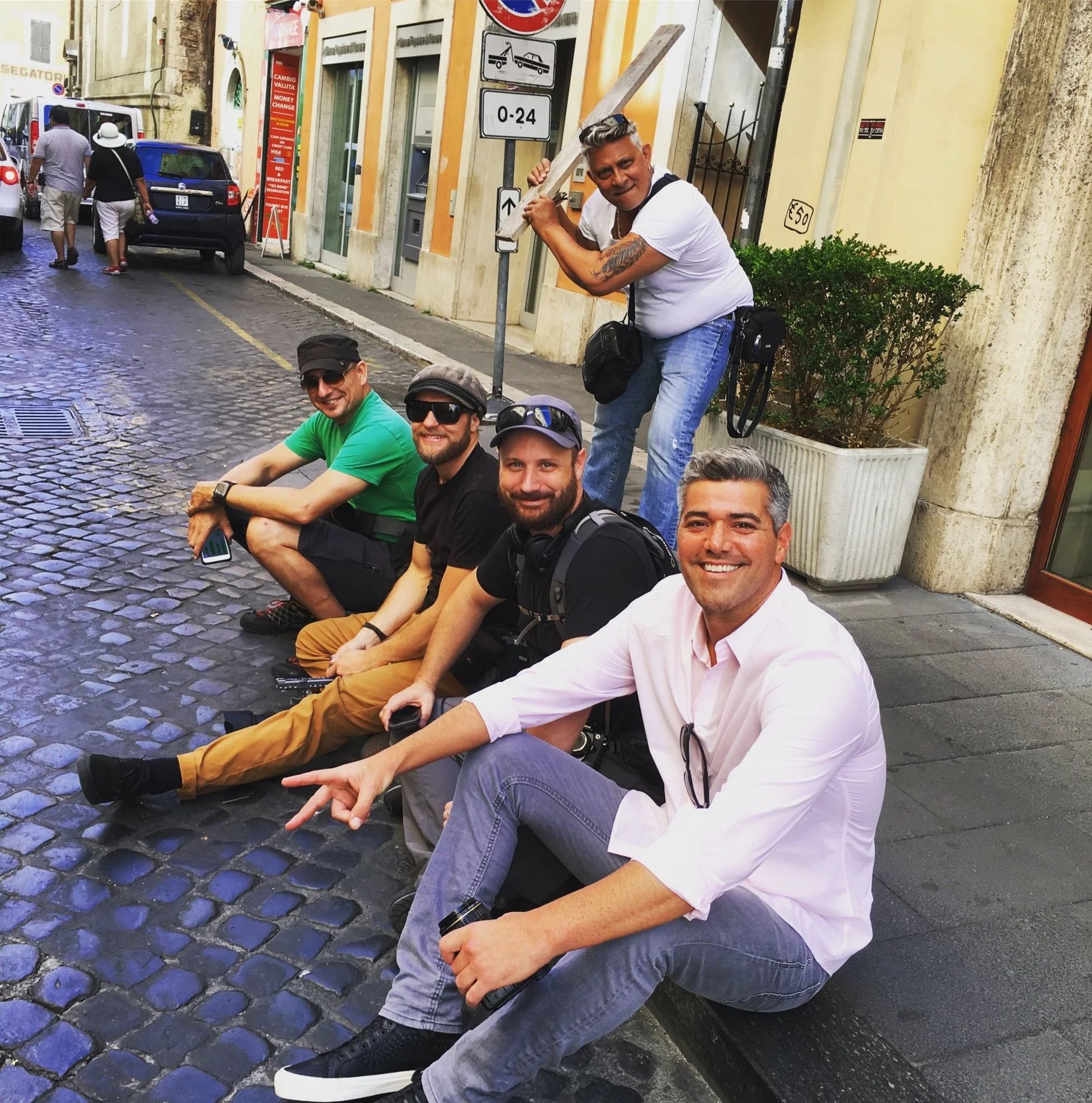 Group of people sitting on cobblestone street in a European city. One man stands behind holding a piece of wood over his shoulder. Others are smiling and looking at the camera. Background includes parked cars, a building with a sign, and street signs.