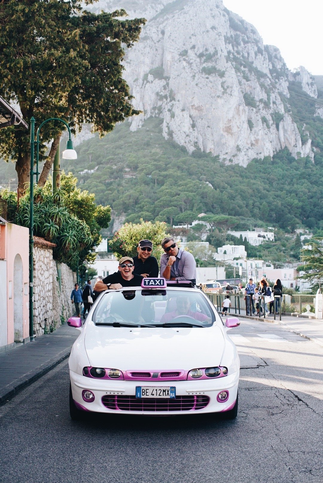 Four men are smiling and having fun on a white taxi with pink accents and a website sticker on the front, parked on a street with a mountain in the background. The photo was taken during the day, and there are other people walking in the background.