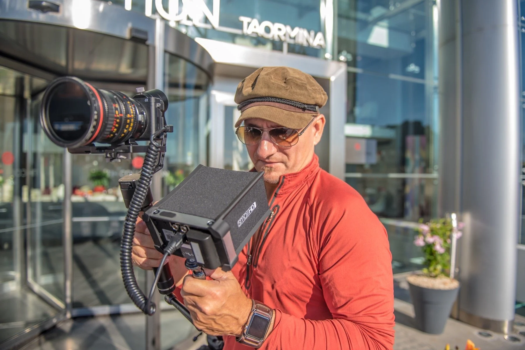 A man with a brown cap, sunglasses, and a red jacket operating a large camera or filming device outdoors in front of a modern building with glass doors and signage.