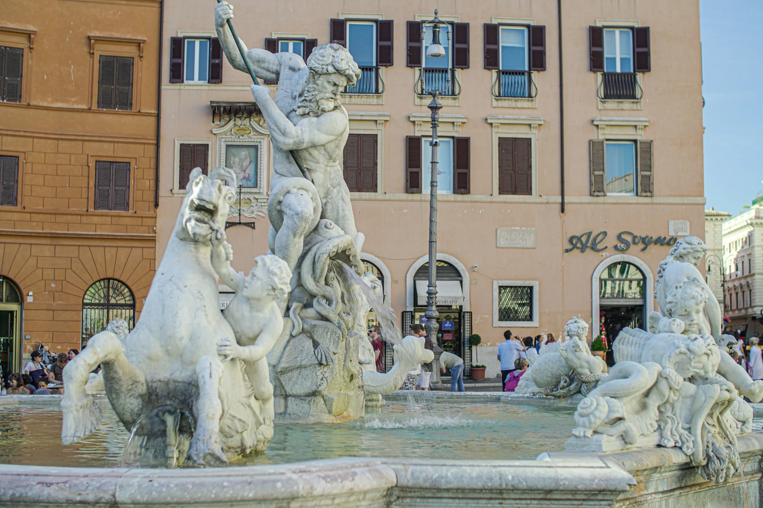 Marble fountain with sculptures of mythological sea creatures and a muscular figure holding a spear, in a busy city square with historic buildings and people walking around.