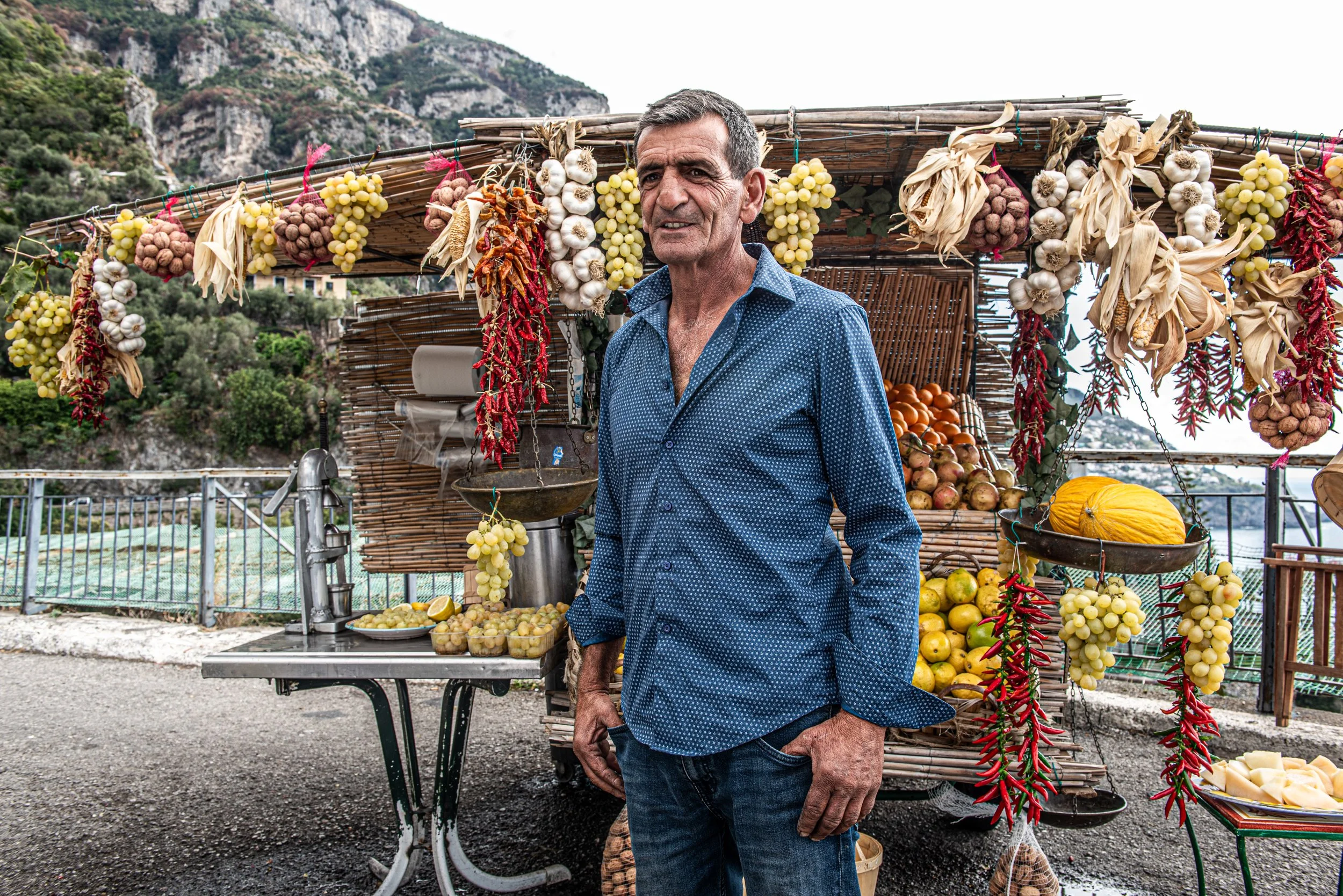 A man stands in front of a stand with hanging and stacked bunches of garlic, grapes, tomatoes, and chili peppers at an outdoor market with mountainous terrain in the background.