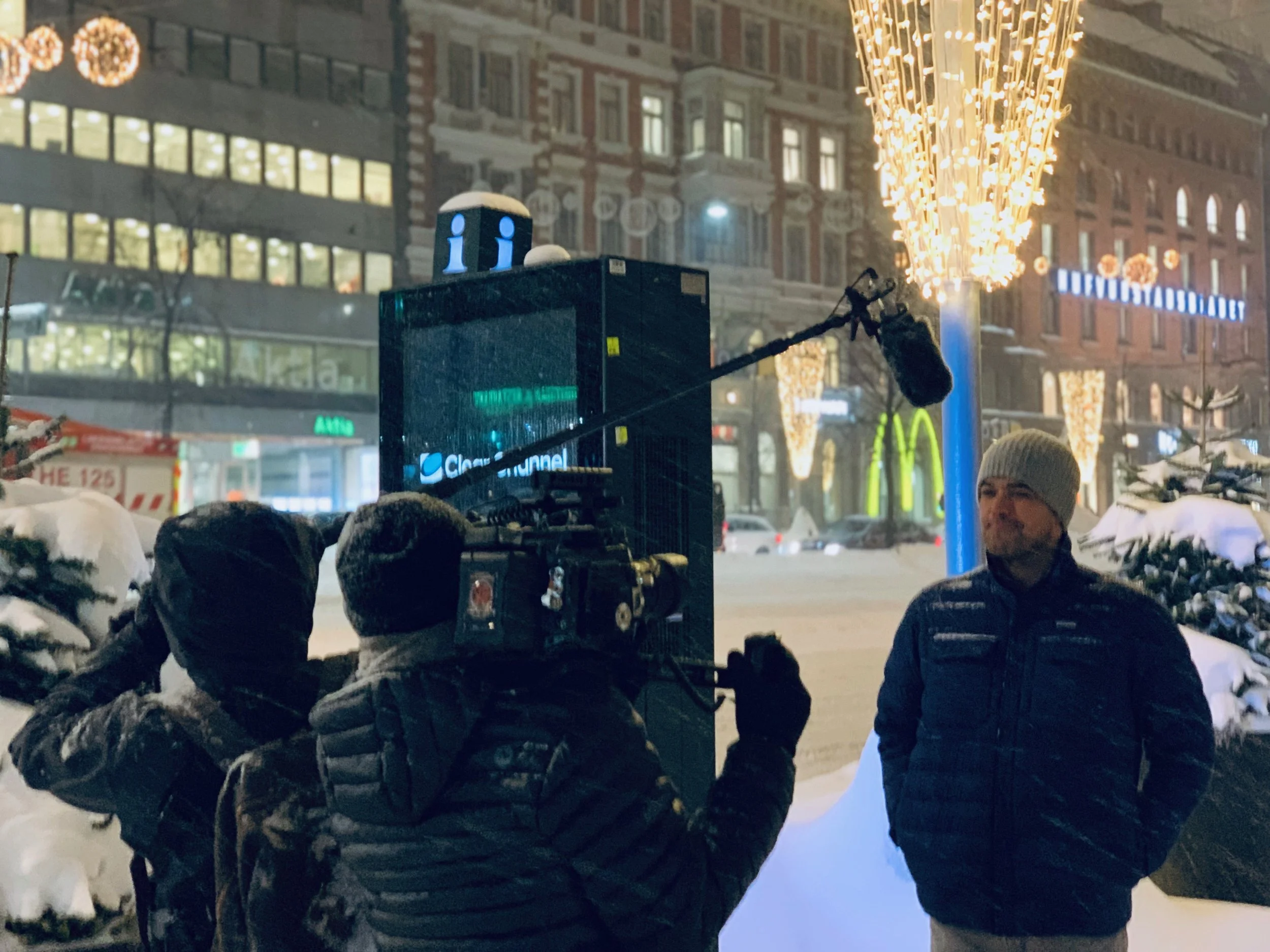 Nighttime scene on a city street with snow, illuminated decorative lights, a crowd of people wearing winter clothing, and buildings in the background.