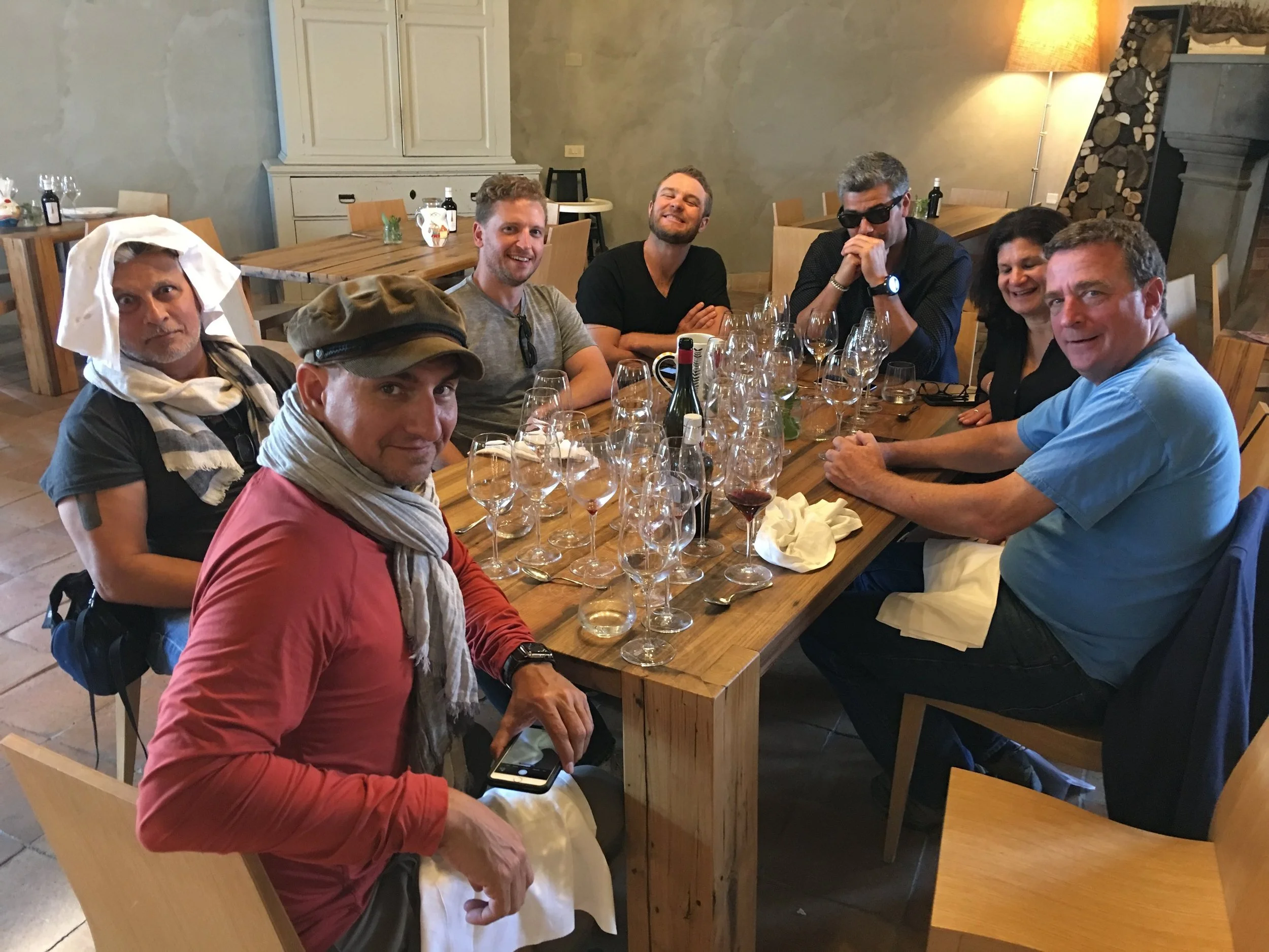 A group of seven people sitting at a wooden dining table in a restaurant, smiling and enjoying each other's company. Some glasses and bottles are visible on the table, and a woman is wearing sunglasses. A man in the foreground is wearing a brown cap and a scarf around his neck.