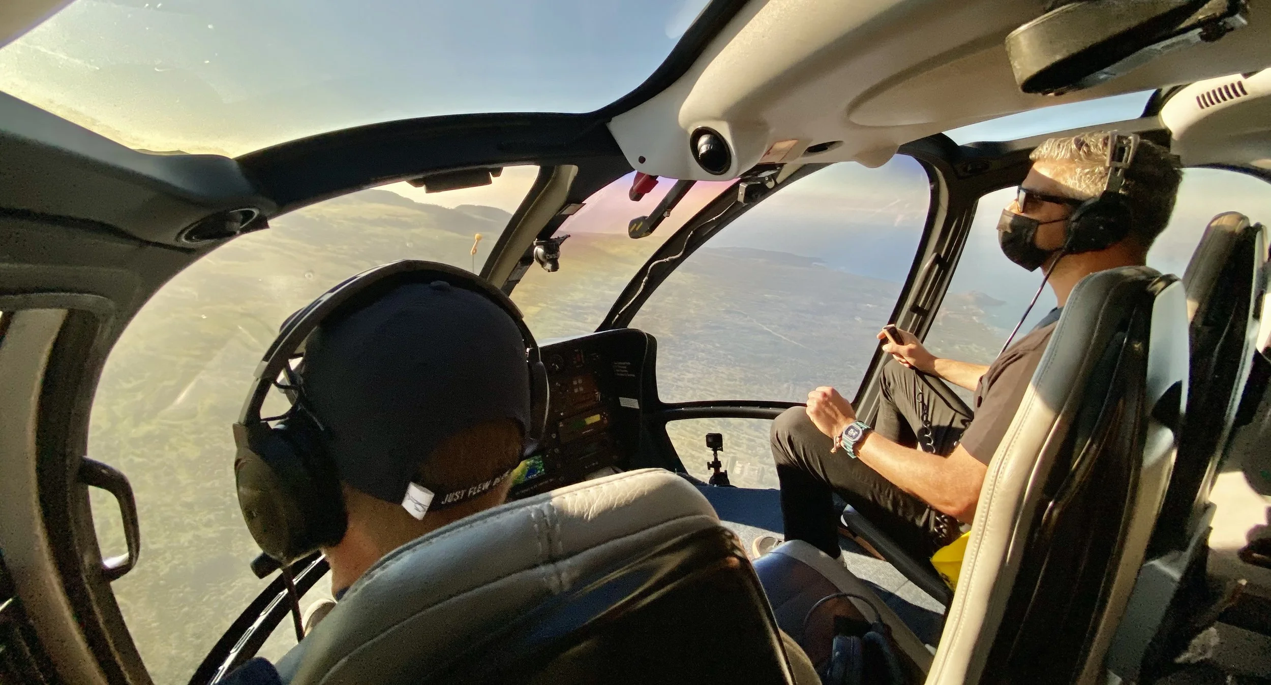 Inside a helicopter cockpit with two pilots flying over a landscape with water, land, and mountains during sunset, one pilot is wearing sunglasses and a black face mask, and the other is wearing headphones and a black cap.
